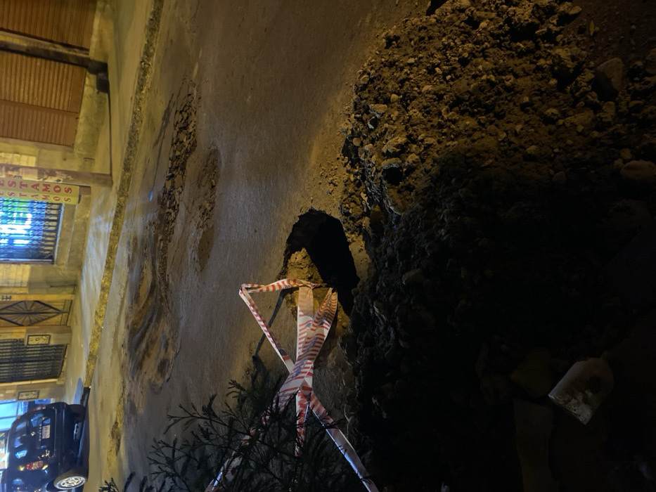 This nighttime image captures an urban street in La Paz, Bolivia, focused on an exposed utility cut or sinkhole. In the immediate foreground, a substantial mound of dark soil and aggregate is piled on the roadside. Adjacent to this, an irregular, dark excavation cuts into the asphalt, partially cordoned off by loose red and white striped barrier tape. Dark green foliage extends into the lower-left corner, partially obscuring the tape.
The asphalt roadway beyond the excavation exhibits significant degradation, marked by multiple potholes, cracks, and uneven sections. A dark-colored Dacia Duster SUV is parked on the street in the mid-ground, facing away from the viewer.
Behind the vehicle, two buildings are visible. The structure on the left features a light-colored facade with a dark, ornate double door. The building to the right has a closed, horizontally-slatted metal roll-down door and a brightly illuminated window displaying partial neon text, including letters like "S," "E," "A," "M," and "O," indicative of a business sign. A wooden utility pole stands vertically on the far right. No individuals are present in the scene.