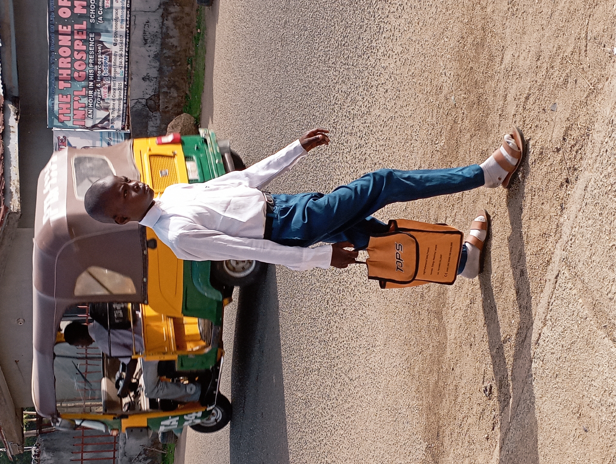A young man stands upright on what appears to be the shoulder of a paved road, next to a sandy or gravel-covered area, in a bright, sunny outdoor setting. He is wearing a long-sleeved white shirt, blue trousers, and light-colored sandals with white socks. He holds an orange-brown bag with the word "TOPS" prominently displayed on it, along with some smaller text underneath. His gaze is directed slightly upwards and to his right, with a neutral expression.
To his left, a yellow and green auto-rickshaw (keke napep), with a dark brown canopy, is parked or moving slowly. A person is visible in the driver's seat. Beyond the vehicle, a large, weathered banner is affixed to a wall or structure. The banner advertises "THE THRONE OF INT'L GOSPEL M" and mentions "AN HOUR IN HIS PRESENCE (Prayer & Intercession)" on "WED. 5:00PM." It also lists social media handles (@Dthroneofgrace, @throneofgrace 2025), a Nigerian phone number (+234-704-365-6357), and contact details including "Botmang Street, Adjacent ITF Corpers Lodge, Da," indicating a specific location in Jos, Nigeria. Other partial text visible on the banner includes "Join Us Us Weekly Ad Vibe Brood © LA C", "SCHOO", "A Come", and "thethroneof". The ground surface transitions from asphalt to a rough, pebbly texture, and strong shadows are cast by the man and potentially other unseen objects, suggesting it is daytime, possibly mid-morning or afternoon.