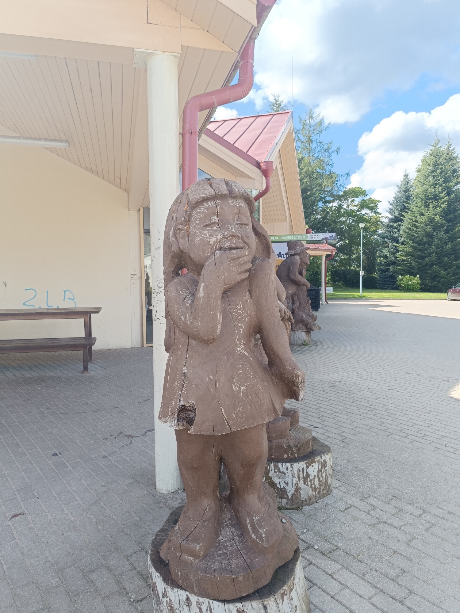 This is a medium shot, eye-level, outdoor, daytime, vertical image of wooden sculptures in the city of Aizpute, Latvia. The sculptures are of folkloric figures. The foreground sculpture is of a young child with hands clasped over its mouth, appearing to be laughing or surprised. The sculpture is made of wood and mounted on a tree stump. Behind it, there are other wooden sculptures of figures in a line, one of which appears to be a woman wearing a hat. A building with a cream-colored facade and a red roof is visible in the background. A light-colored bench with graffiti on the wall behind it is to the left of the frame. The ground is paved with gray interlocking bricks. Evergreen trees are visible behind the building and to the right of the frame. A white pole is to the right of the foreground sculpture. A red car is parked on the right side of the image. The sky is blue with white clouds.
