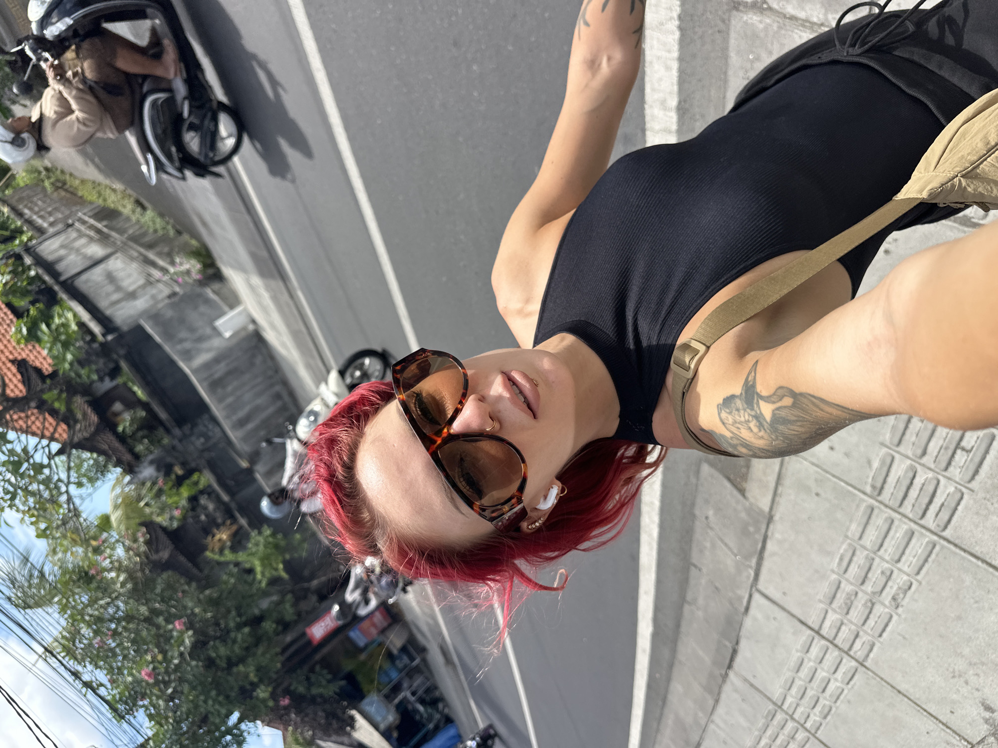 A fair-skinned person with red hair, wearing sunglasses and a black tank top, stands on a concrete sidewalk. They are holding a mobile device, ostensibly taking a selfie. Tattoos are visible on the person's arm. A khaki-colored bag strap crosses their shoulder. The backdrop is a city street in North Kuta, Indonesia, featuring parked scooters, urban greenery, and commercial signage in Indonesian. The street itself is gray asphalt.