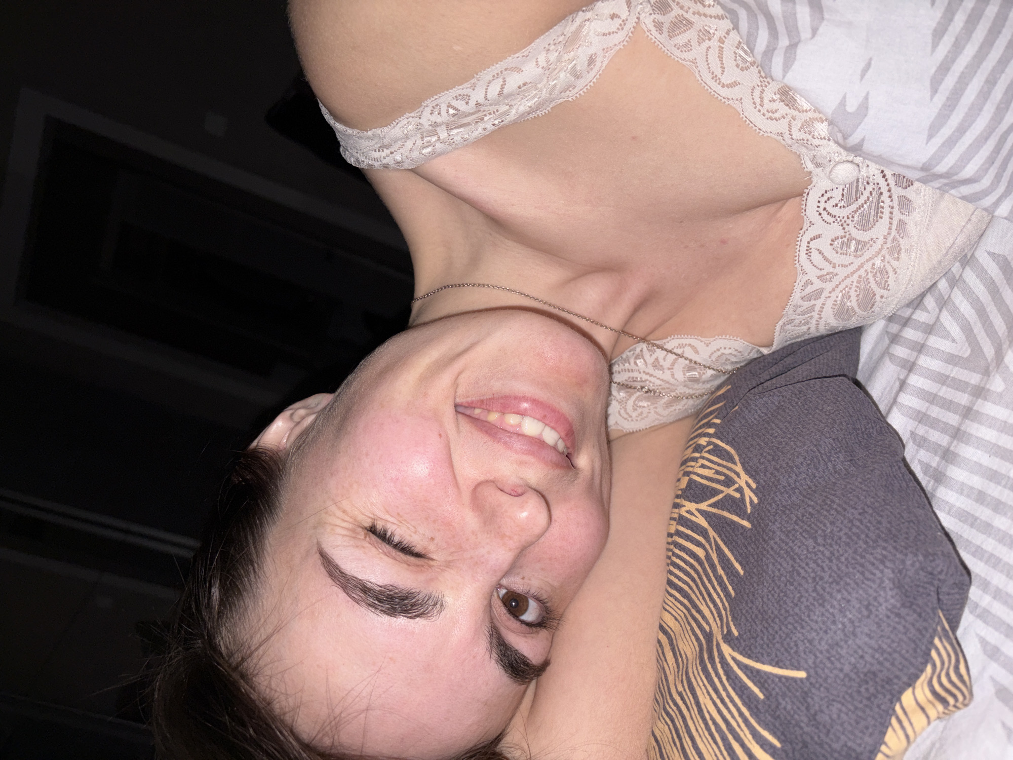 A woman is lying down, tilted towards the left of the frame, smiling and winking with one eye. She has fair skin, dark brown hair pulled back, and full eyebrows. She wears a lacy white camisole and a silver necklace. She appears to be lying on striped bedding, with a gray patterned pillow or blanket. A dark window is visible in the upper left corner.
There is no information to suggest that this image was taken in Izhevsk, Russia.