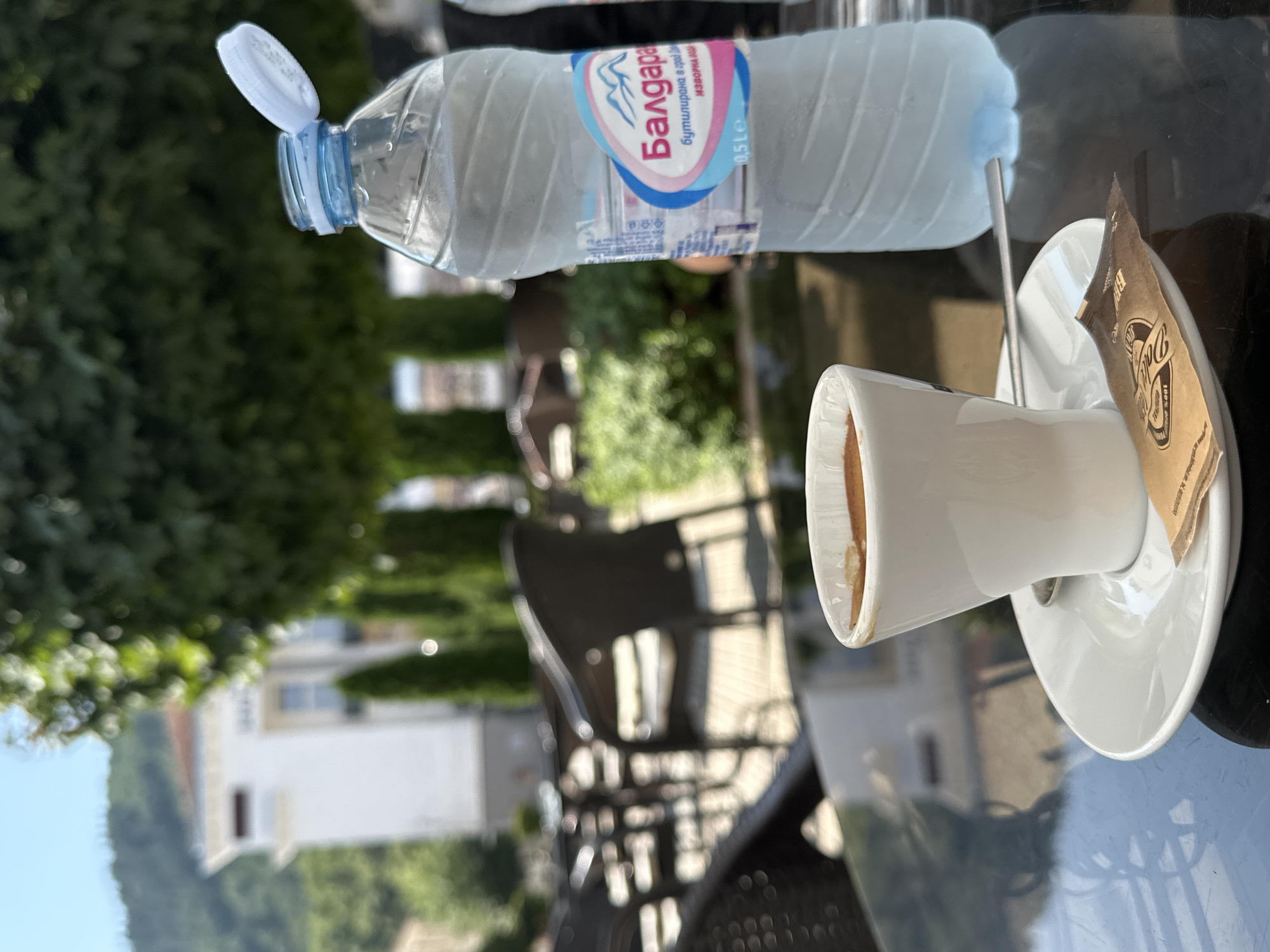 A white ceramic cup filled with dark liquid, likely coffee, sits on a saucer on a dark reflective table. A silver spoon rests on the saucer next to a small brown paper packet. To the right of the cup is a partially filled plastic bottle of water with a blue cap open, its label indicating it is from "Baldara" mineral water, with the text "бутилирана в гр. Петрич, България" (bottled in Petrich, Bulgaria) visible. In the background, the scene suggests an outdoor cafe setting with dark wicker chairs and tables visible. Trees and a building with a white facade and red-tiled roof are present. The overall environment appears to be a sunny day in the city of Petrich, Bulgaria. There are no people clearly visible in the frame.