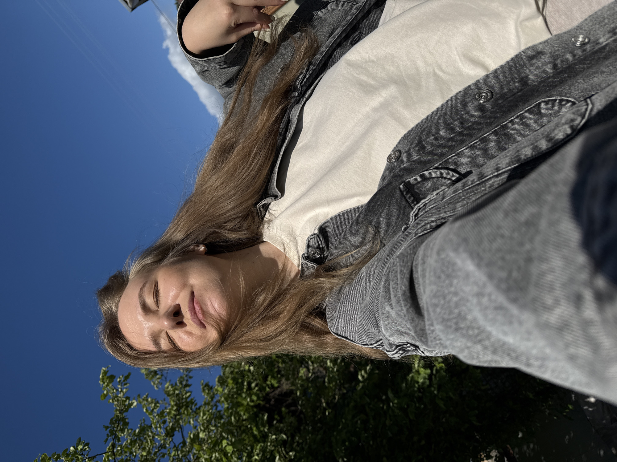 A selfie captures a young person, likely female, outdoors in Красный Маяк, Russia. Her head is positioned in the lower part of the frame, tilted slightly upwards towards a clear, vibrant blue sky which dominates the upper portion of the image. Her eyes are gently closed or squinting, and a soft, contented smile plays on her lips, suggesting enjoyment of the bright sunlight. Long, light brown hair flows down, framing her face. She is dressed in a light-colored (possibly off-white) t-shirt worn under a dark grey denim jacket, which features metallic snap buttons. The natural lighting highlights her facial features, creating a subtle sheen on her forehead and lips. In the bottom left corner and along the bottom edge, lush green tree foliage is visible, indicating a natural, outdoor setting, possibly a park or garden. The image conveys a serene and peaceful mood, reflecting a moment of calm under a sunny sky. There is no visible text in the image.