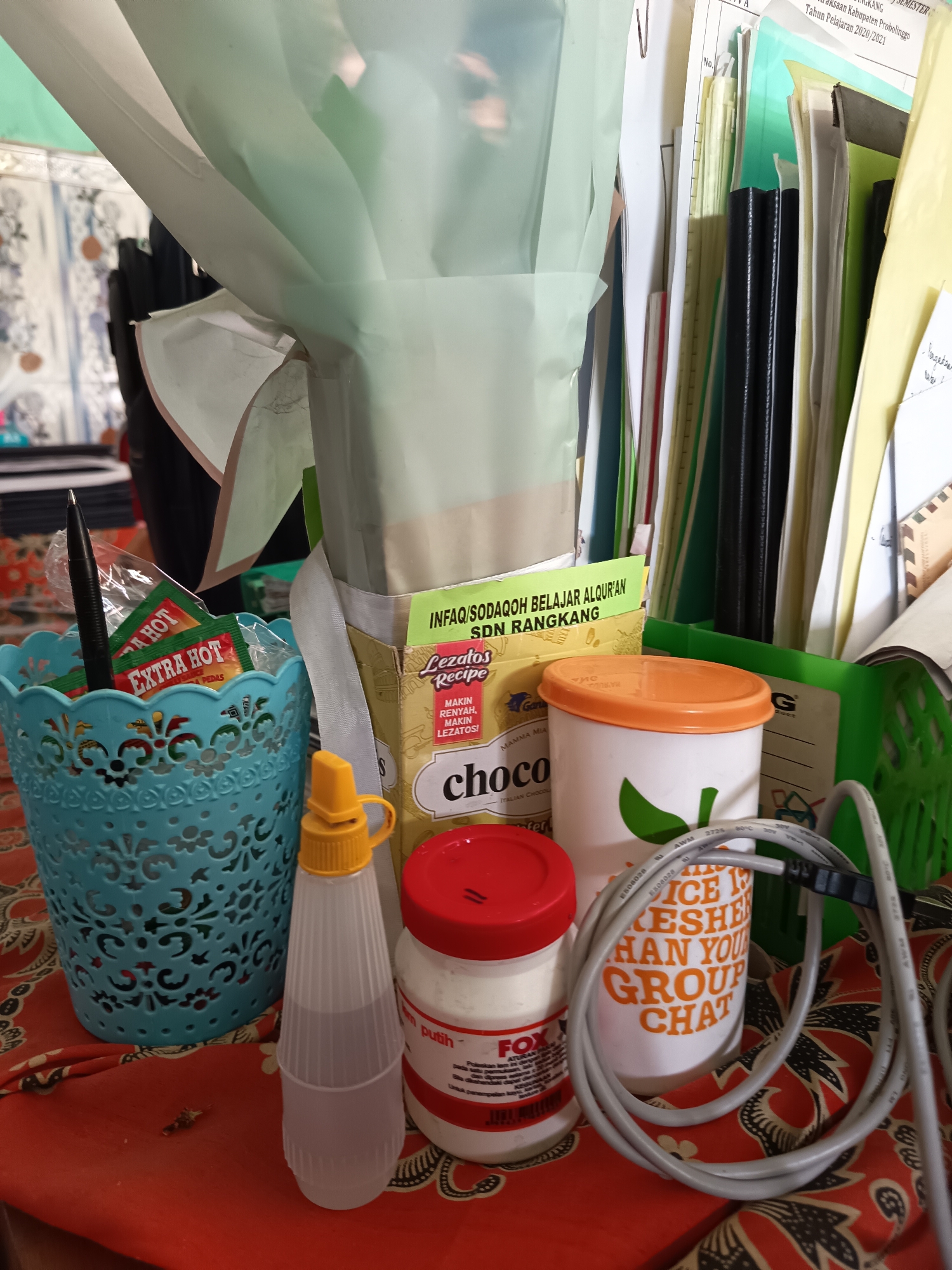 In a close-up, overhead shot, a collection of items is arranged on a patterned orange cloth. On the left, a teal filigree basket holds a black pen and several small packets labeled "EXTRA HOT" and "EXTRA HOT SAUCE". To the right of the basket is a clear plastic squeeze bottle with a yellow cap. In the center, a cardboard box with green and yellow text reads "INFSO/SODAQOH BELAJAR AL-QUR'AN SDN RANGKANG" and "Lezatós RECIPE MAKIN RENYAH MAKIN LEZATOS!". Below this box is a white container with an orange lid, featuring an image of a green leaf and text that reads "ICE IS KESHER HAN YOU GROUP CHAT". Next to it is a white jar with a red lid and branding that includes the word "FOX". In the background, stacks of papers and binders are visible, some with Indonesian text. The overall scene appears to be an indoor arrangement, possibly a desk or table, in Widoro, Indonesia, given the context.
