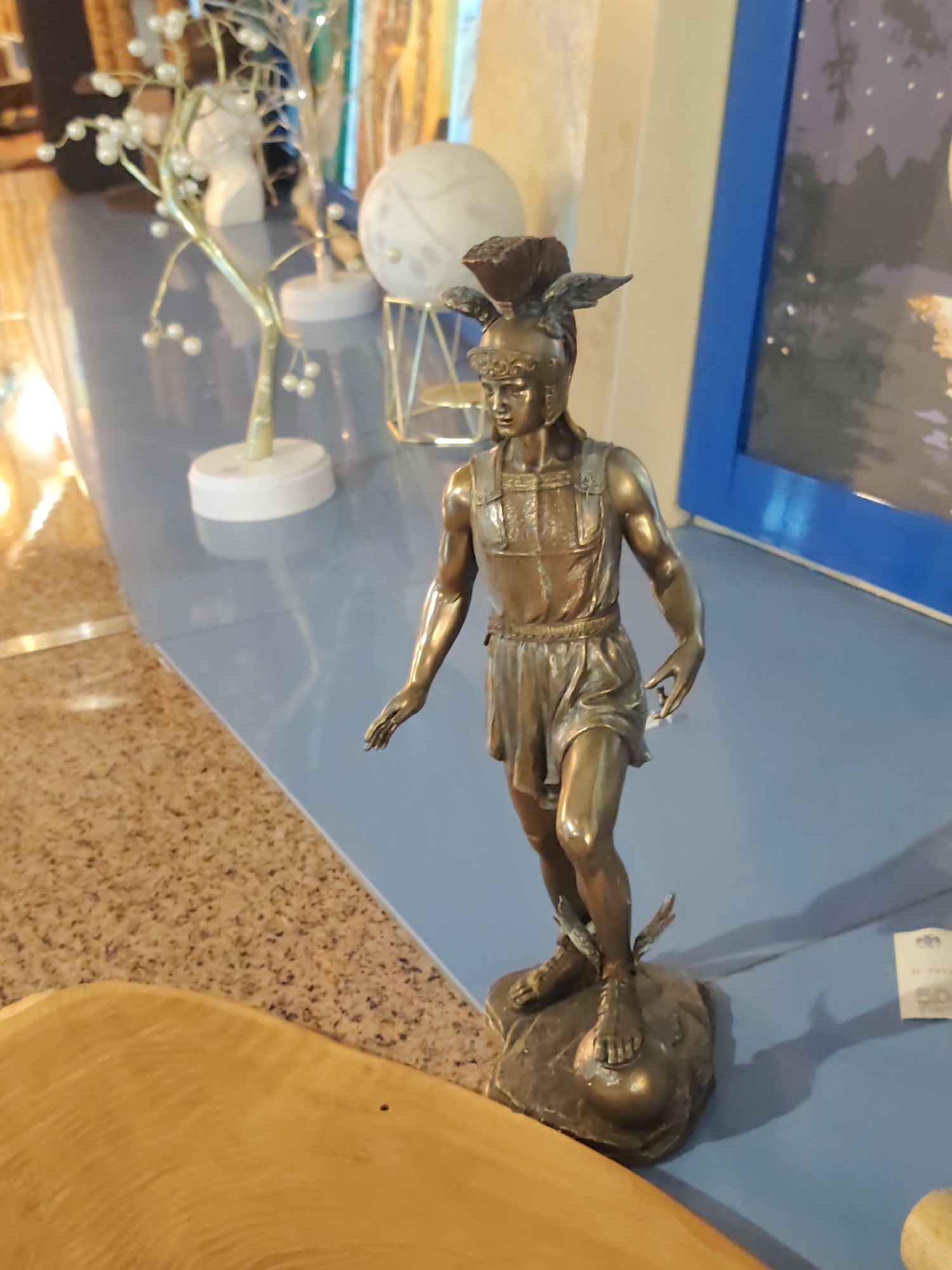 This image shows a bronze statue of a winged figure, likely Mercury or Hermes, standing on a rock with a sphere beneath its feet. The statue is detailed, with the figure wearing a helmet with wings, a tunic, and sandals with wings. The background includes decorative elements like a tree-like object with pearl-like embellishments and a framed artwork with a nighttime scene. The setting appears to be an indoor display or shop. Based on visual clues that might indicate the location, such as the style of decor and potential signage (though unreadable), it's impossible to definitively place this scene in Sochi, Russia.