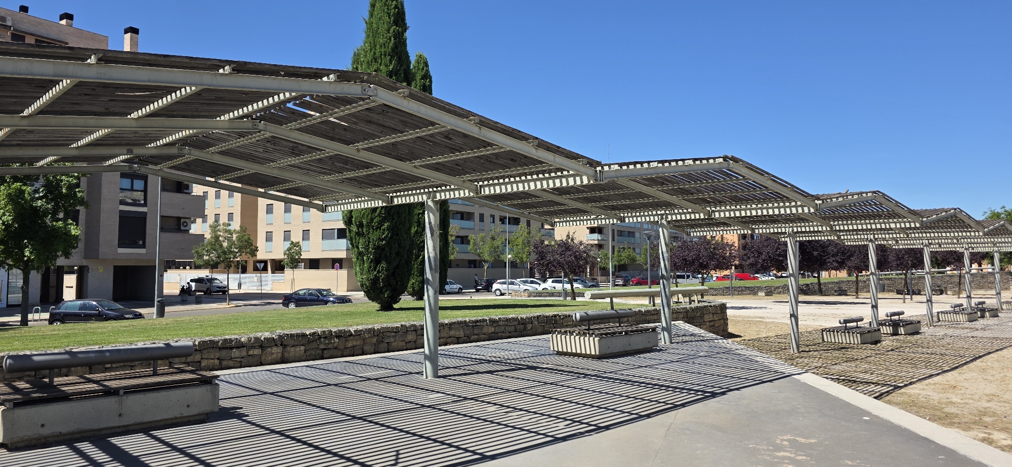 This image depicts a park scene under a bright, clear blue sky in Huesca, Spain. In the foreground, a row of modern pergolas with slatted wooden roofs provides shade over a paved area. Underneath the pergolas are several concrete benches with metal seating. A stone wall borders a grassy area behind the pergolas. Across the grassy area, a street is visible with several parked cars and some passing vehicles. In the background, apartment buildings of varying heights and architectural styles line the street, interspersed with trees. Tall, slender cypress trees and trees with dark purple foliage are prominent features of the landscape. The overall impression is of a well-maintained urban park designed for public use, likely on a sunny day.