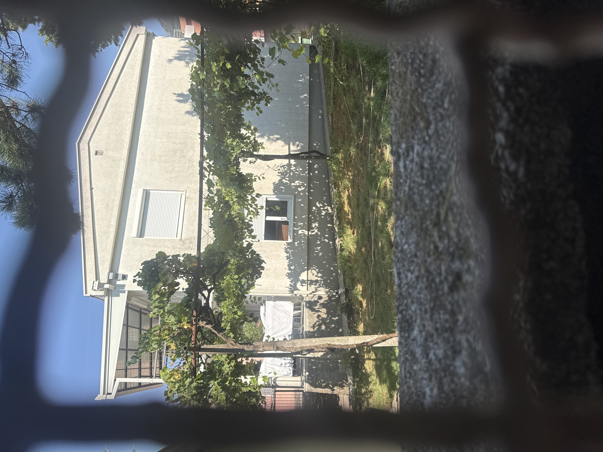The image shows a residential building with a light-colored facade, a sloped roof, and a covered patio area. In front of the building, there is a garden with a trellised vine. Laundry is hanging on a clothesline in the background. The view is partially obstructed by a blurred foreground element, possibly a fence or bars. The scene appears to be in a suburban or residential area under a clear blue sky. The location is Zgrade, Montenegro.