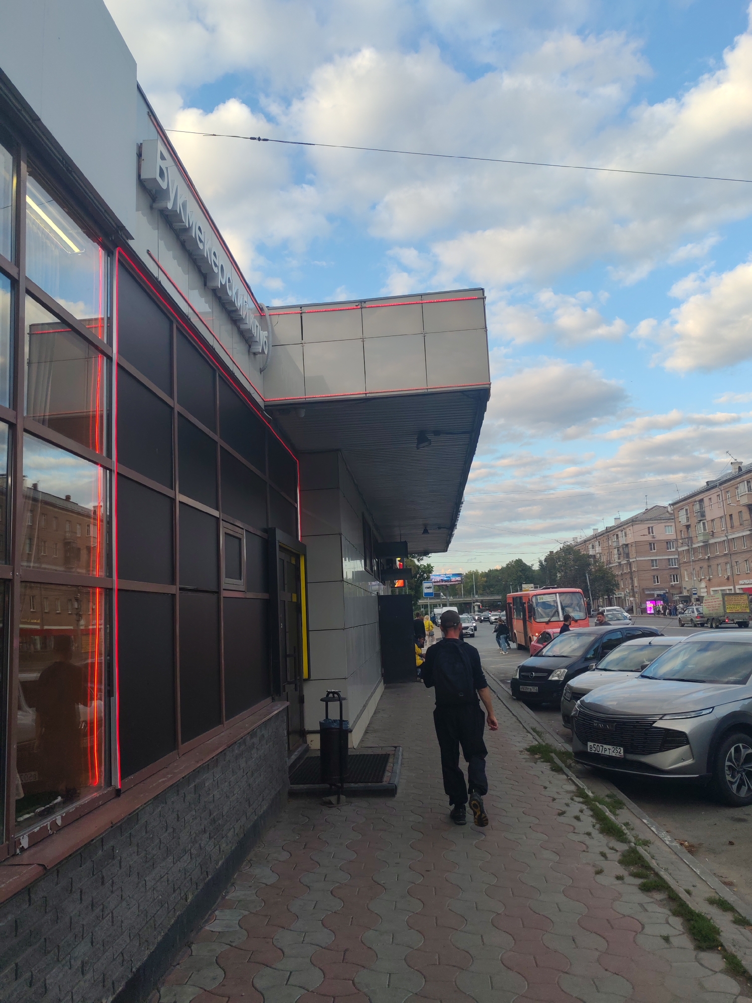 In Nizhny Novgorod, Russia, on a cloudy day, a man wearing a black cap, black t-shirt, and dark pants with a backpack walks along a sidewalk next to a building with large windows and red neon trim. The building has signage visible that appears to be in Cyrillic. Across the street, there is a red bus, several cars, and apartment buildings. The street is paved with patterned interlocking concrete pavers. The scene appears to be a street-level view of a commercial or public area.