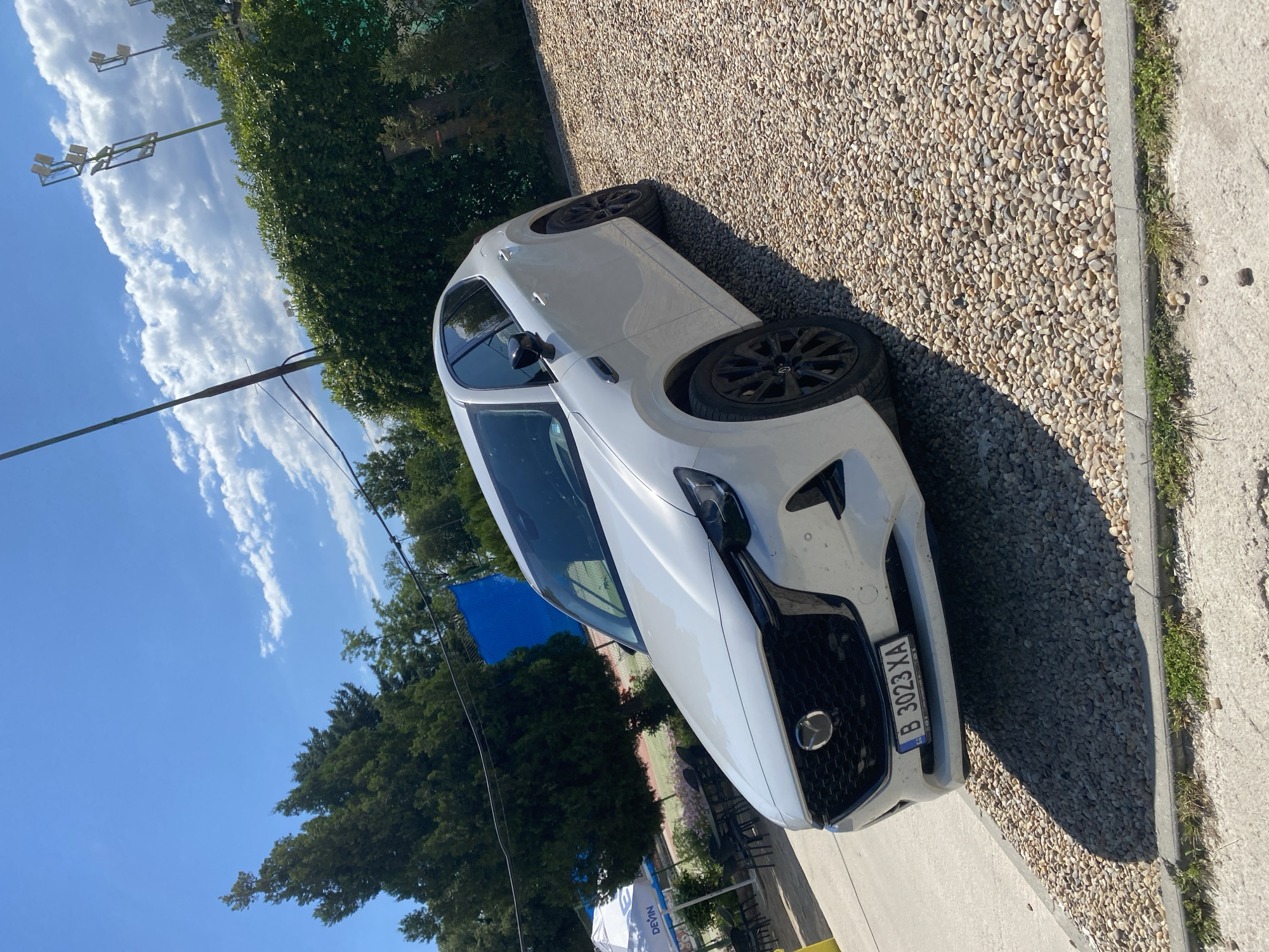 The image shows a white Mazda CX-5 parked on a gravel surface next to a concrete path. The car is facing towards the right side of the frame. The front of the car, including the grille and headlights, is visible. A license plate with the text "B 3023 XA" is attached to the front bumper.
In the background, there are lush green trees and some sports facilities, possibly a tennis court, indicated by a blue tarp. The sky is a clear blue with some wispy white clouds. The sun appears to be high in the sky, suggesting it is daytime. The overall scene appears to be outdoors in a sunny and pleasant environment, likely a park or recreational area, possibly in Varna, Bulgaria, as indicated by the location context. There are no people visible in the image. The focus is on the car and its surroundings.