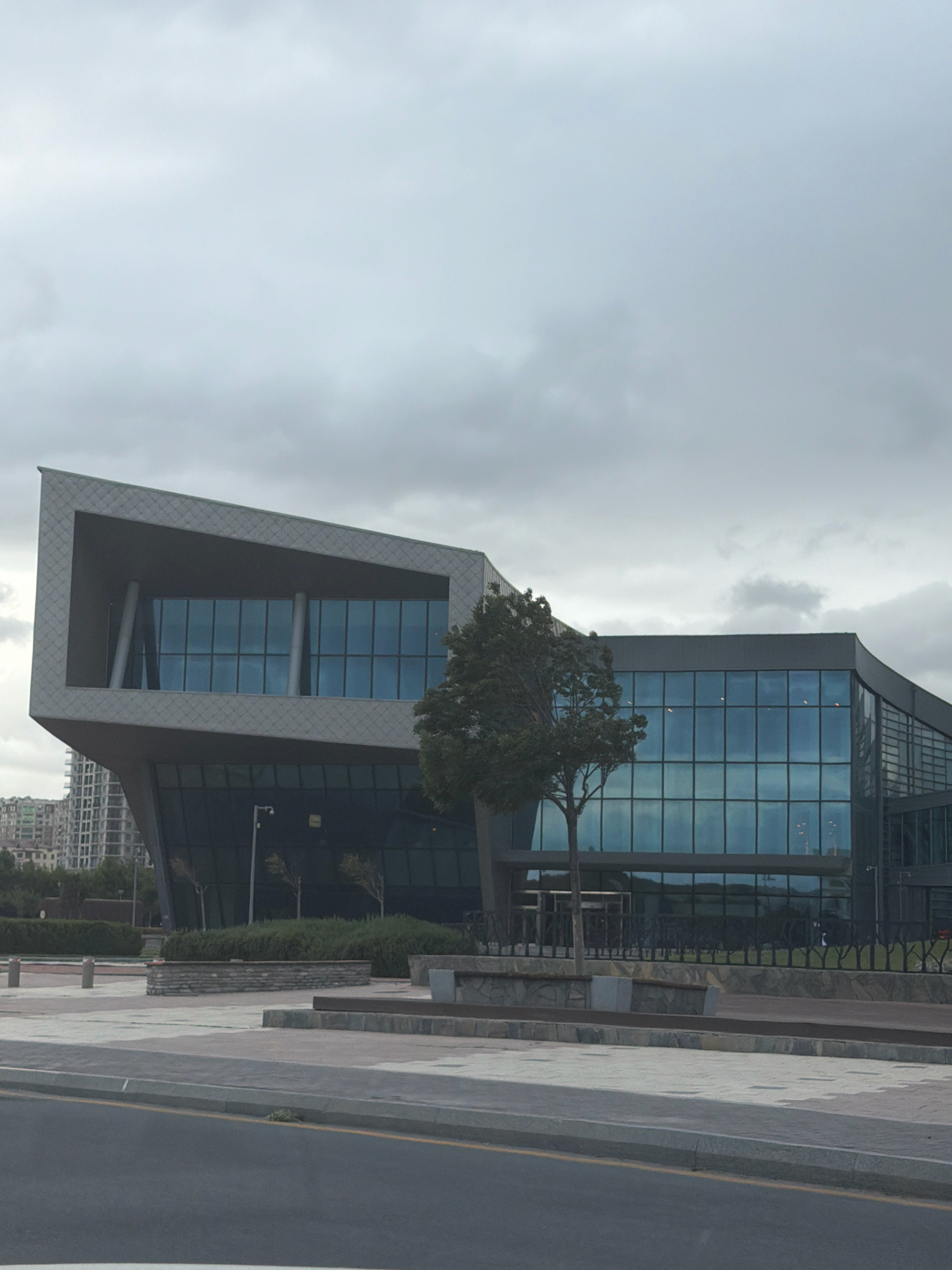 The image depicts a modern architectural building in Baku, Azerbaijan, under an overcast sky. The building features a striking geometric design with large glass windows and angled facades. A lone tree stands in front of the glass-paneled section of the building, and there is landscaping with low hedges and stone elements. A sidewalk and a road with yellow and grey markings are visible in the foreground. In the background, other buildings, including high-rises, can be seen. The overall scene suggests an urban environment with contemporary architecture.