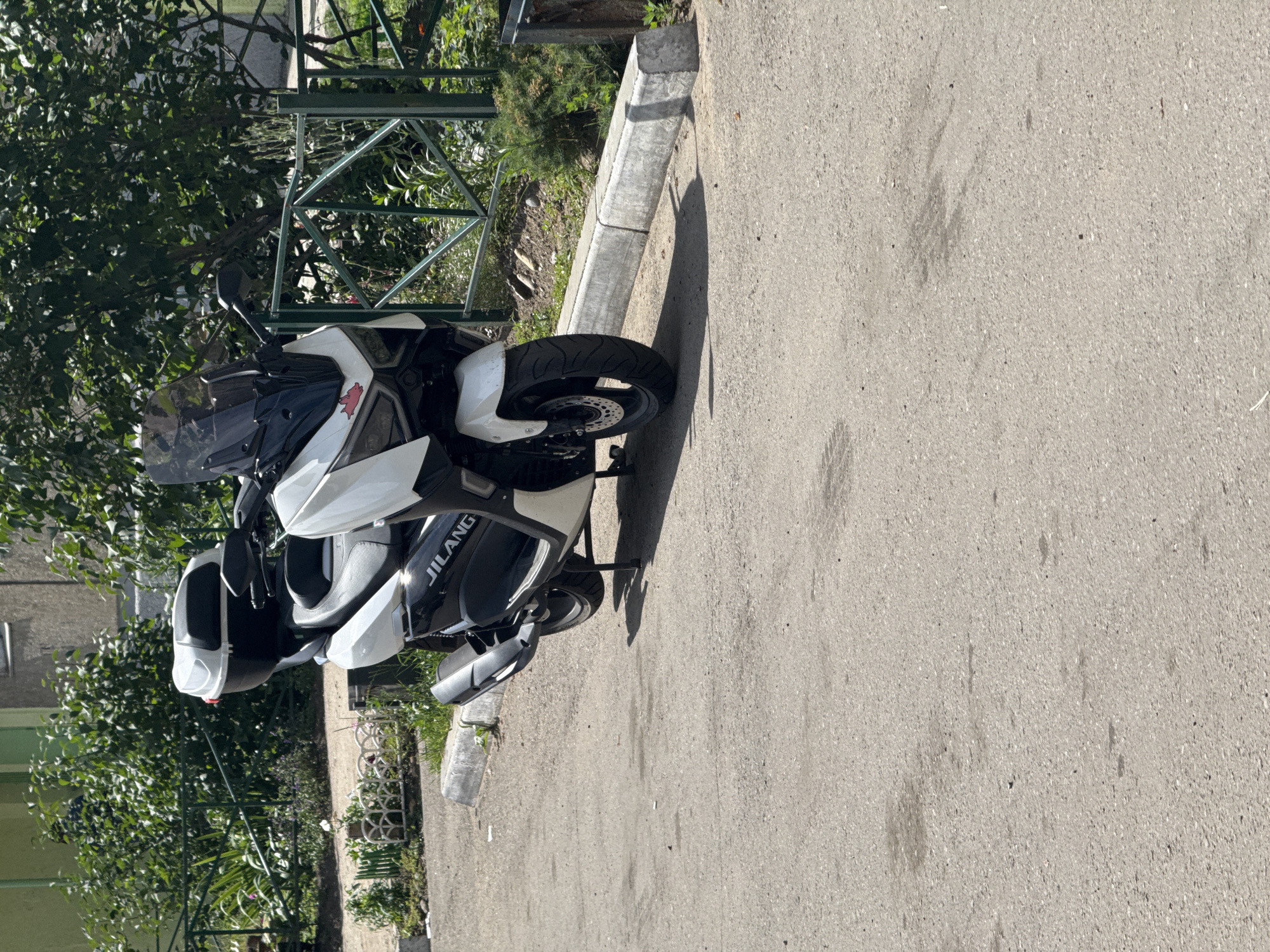 A white and black JILANG scooter is parked on an asphalt surface next to a concrete curb. The scooter has a large black top case and a tinted windshield. Behind the scooter, there is a green metal fence and lush green foliage. In the background, a building with a green facade is partially visible. The scene is illuminated by daylight. The location is the city of Angarsk, Russia.