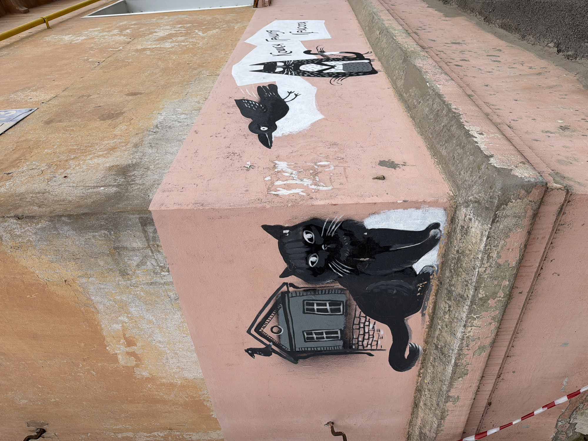 A low-angle, angled shot captures street art in Votkinsk, Russia. The art is painted on the side of a building or structure, featuring black and white illustrations on a pink-toned surface. The artwork includes a black cat with a house underneath, a crow, and another cat-like creature with text above it. The overall style appears whimsical and illustrative. The surrounding architecture shows signs of age and weathering.