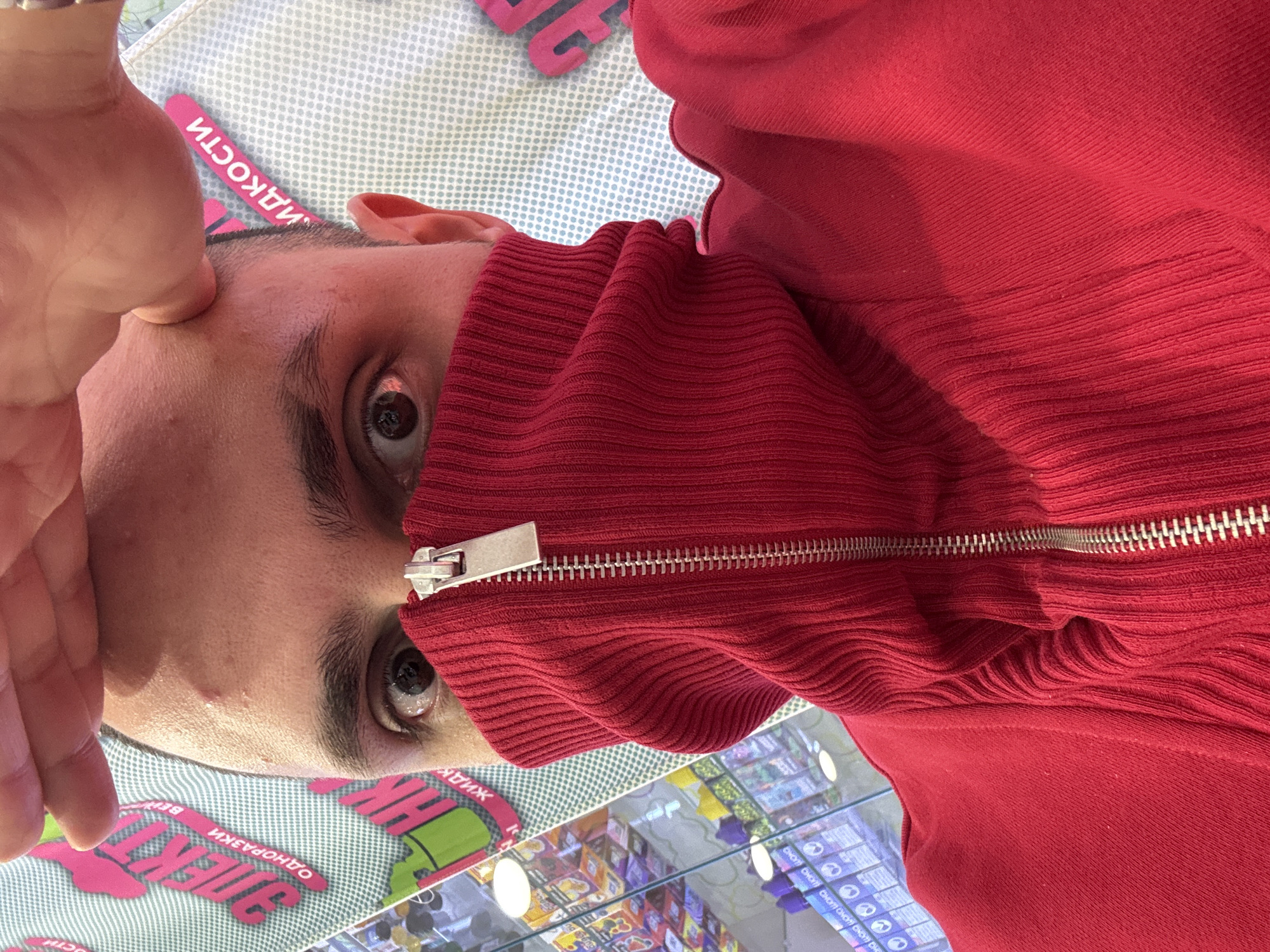 A man wearing a red zip-up sweater partially covering his face is shown in a selfie. His left hand is raised to his forehead. The background shows a glass display case filled with various items in packaging, indicating a store or retail setting. Russian text and brand names are visible on the packaging in the display case. The location is likely within a commercial area in Saint Petersburg, Russia based on the product packaging.