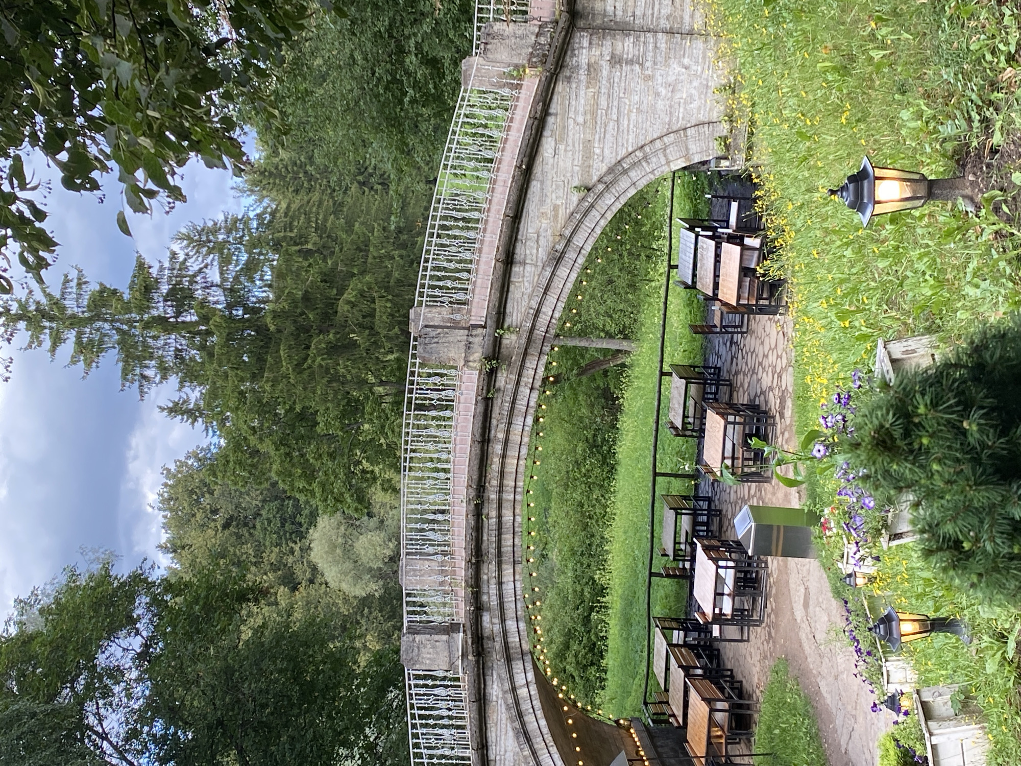The image depicts an outdoor seating area under a stone bridge, likely a restaurant or cafe, located in the city of Pavlovsk, Russia. The bridge, made of light-colored stone blocks, features an ornate white railing along its top and is illuminated by a string of yellow lights beneath its arch. The area beneath the bridge is paved and furnished with multiple wooden tables and chairs, some of which are arranged in small groups.
The environment surrounding the seating area is lush and green. A variety of trees, including tall coniferous trees and deciduous trees with full green foliage, form a dense forest backdrop. The ground is covered in grass and dotted with numerous small yellow flowers, creating a vibrant meadow-like appearance. Several white planters containing purple flowers are visible in the foreground, along with two black lampposts emitting a warm light. A modern, metallic trash receptacle stands near some of the tables. The sky above is cloudy, with patches of blue visible. No people are present in the scene.