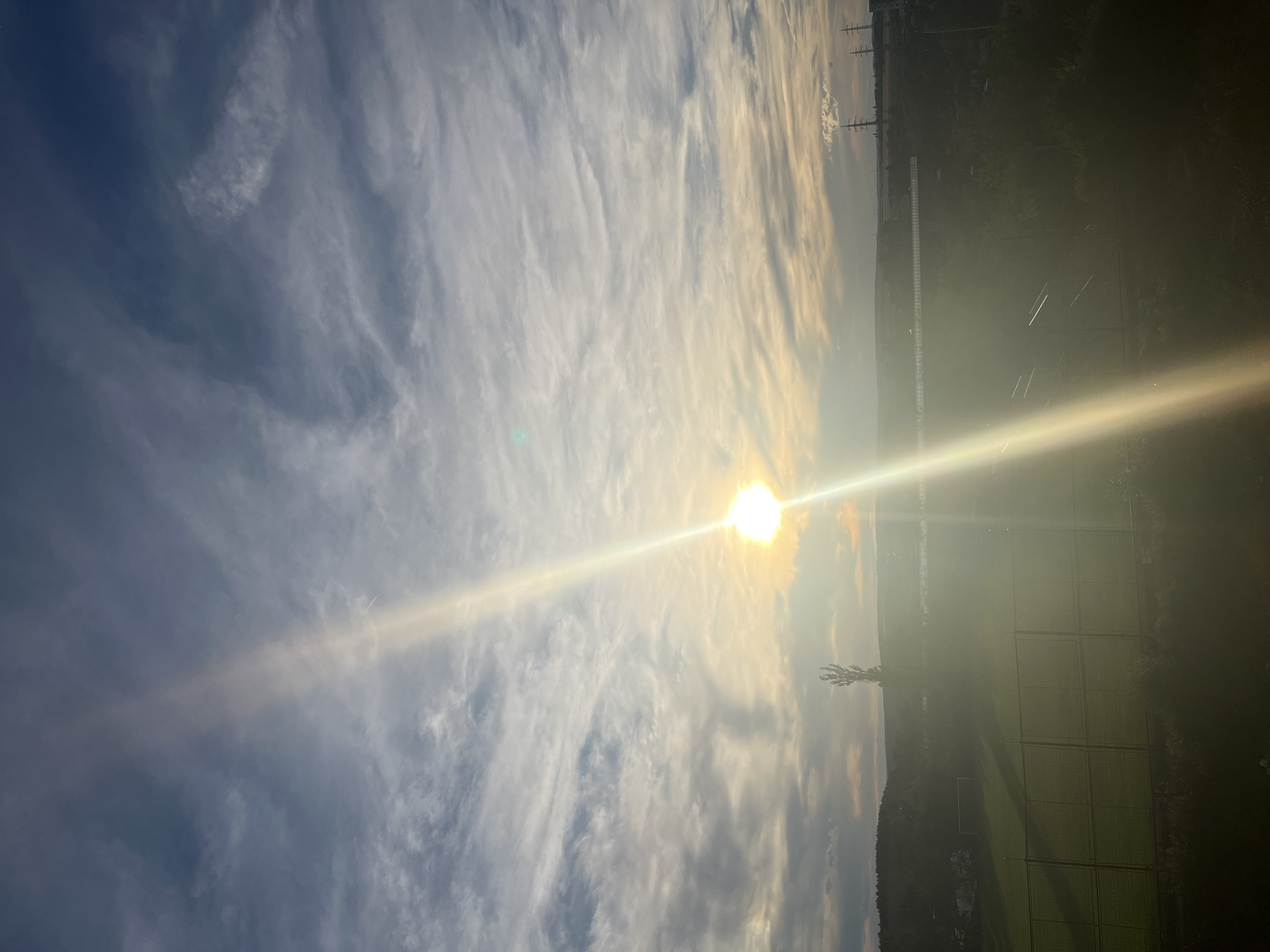 A wide shot captures the sky over Pilsen, Czechia, at sunset. The sun, a bright orb, is partially obscured by clouds and emits a strong lens flare that dominates the center of the frame. Wispy, white clouds stretch across a deep blue sky. In the distance, a silhouette of a tree stands tall on a hill. Below, a green sports field with a goal is visible, bordered by a fence. Power lines and buildings can be seen further in the background, suggesting an urban or suburban environment. The lighting indicates it is either sunrise or sunset.