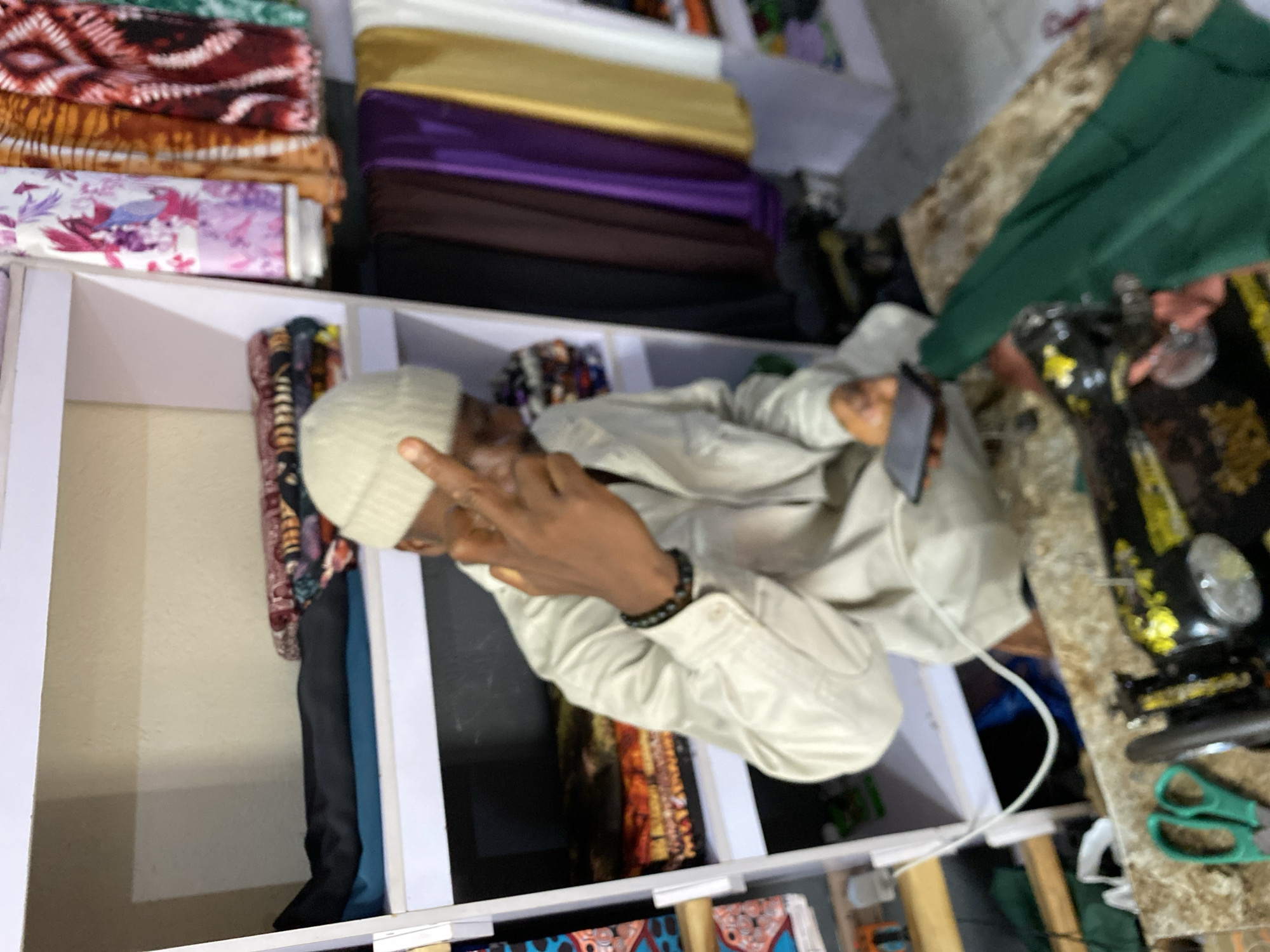 A person wearing a light-colored shirt and a white knit cap is in what appears to be a tailor's shop or a fabric store in Bukuru, Nigeria. The person is making a peace sign with their right hand and holding a smartphone in their left hand. The individual stands beside a white shelving unit holding rolls of colorful fabric. A sewing machine is nearby, sitting on a surface strewn with materials and sewing tools.