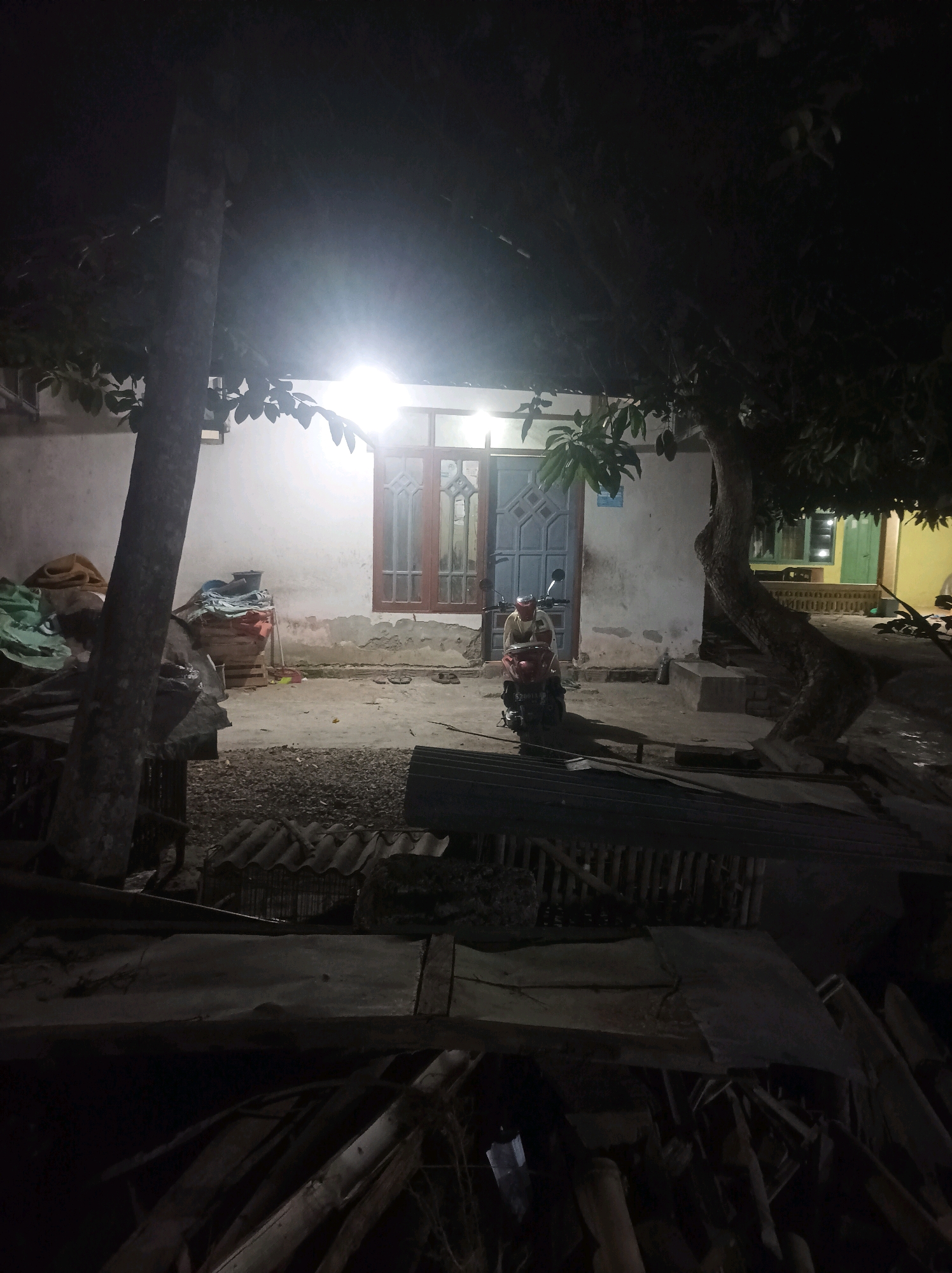 The image depicts a dimly lit outdoor scene at night in Canga'an, Indonesia. In the foreground, there are piles of lumber and debris. The midground features a light-colored house with a window on the left and a blue double door on the right, both illuminated by an overhead light. A person is seated on a red scooter in front of the house, facing left. To the left of the house, a large tree trunk with branches and leaves is visible. To the right of the house, another large tree with dark foliage is prominent. The ground appears to be a mix of dirt and gravel, with some corrugated metal sheeting in the midground. The overall impression is that of a residential yard at night, possibly in a rural or semi-rural setting.