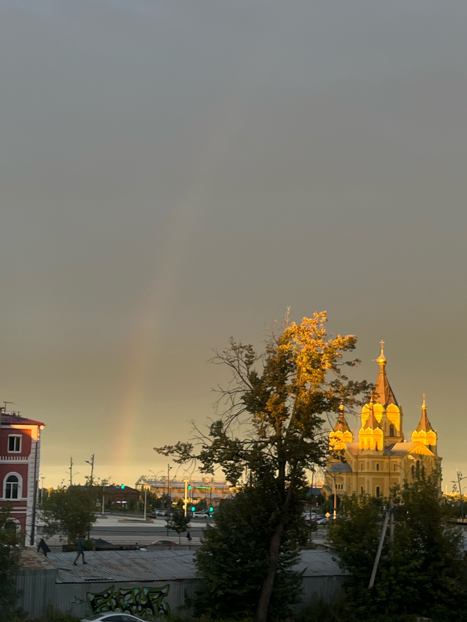 In Nizhny Novgorod, Russia, the image captures a twilight scene with a faint rainbow arcing across a muted, hazy sky. The sun, setting behind heavy clouds, casts a warm, golden light on the architectural splendor of the Alexander Nevsky Cathedral, illuminating its numerous domes and spires. In the foreground, a stark red building with arched windows stands to the left, its upper floors catching the last rays of sunlight. Below, a row of low-lying structures, some with corrugated metal roofs, stretches across the middle ground. Two individuals are visible on one of these rooftops; one, wearing dark clothing, appears to be walking away, while another, in lighter attire, is standing and facing away from the camera. The scene is populated by trees, some bare and skeletal, others lush with foliage, interspersed with streetlights and distant city buildings, suggesting an urban environment. The overall atmosphere is tranquil, marked by the ethereal glow of the setting sun and the subtle presence of the rainbow.