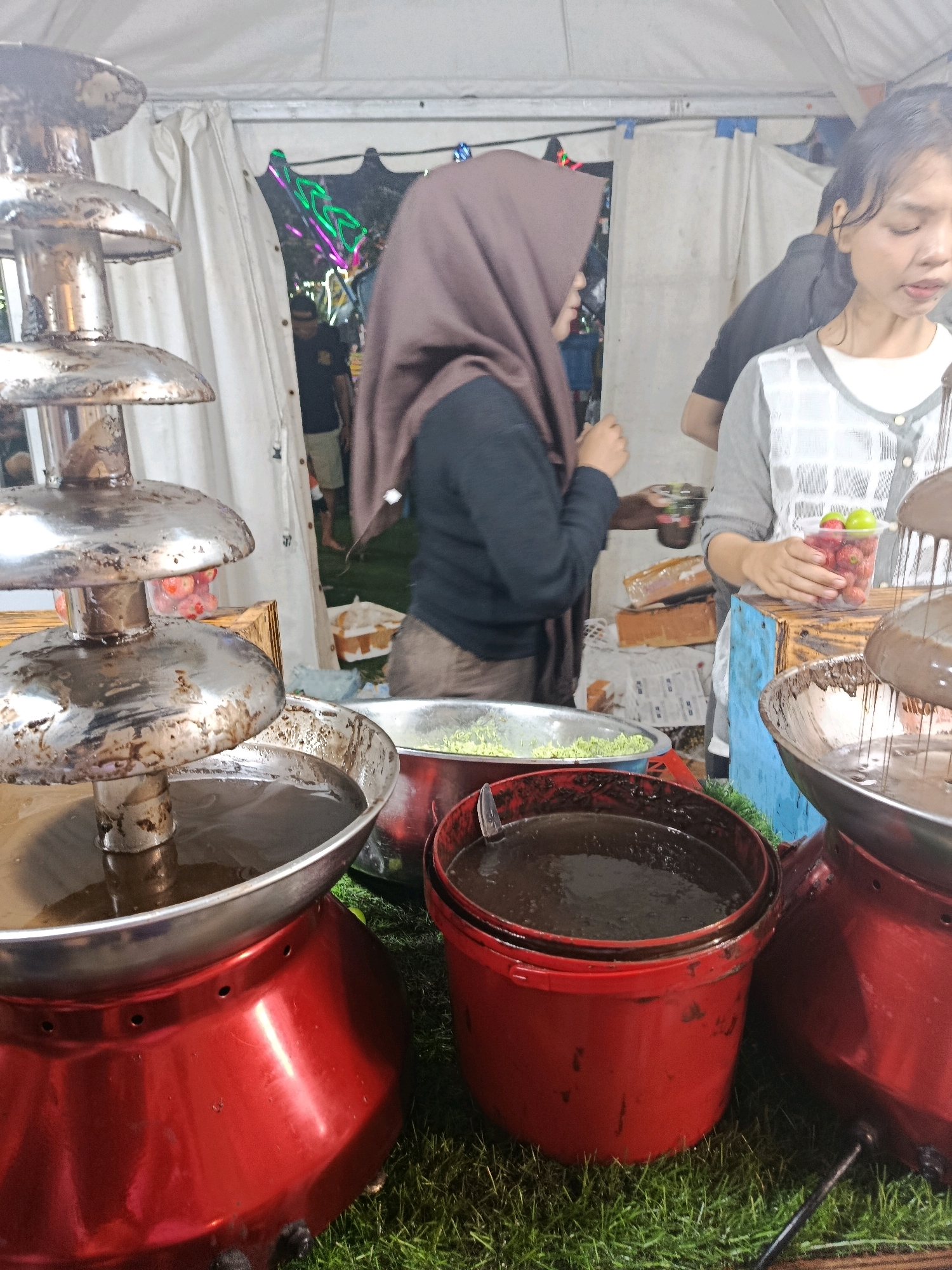 The image depicts a close-up view of a chocolate fountain setup at what appears to be an outdoor event or market stall, likely in Balaraja, Indonesia. In the foreground, a large, multi-tiered stainless steel chocolate fountain with a bright red base is prominent. The fountain is filled with melted chocolate, which is flowing down the tiers. Beside the fountain are two large red buckets, one filled with dark liquid, possibly more melted chocolate or a related ingredient, and another empty or holding a similar dark liquid. To the right of the fountain, a woman wearing a grey and white checkered top and a white apron is holding a clear plastic cup filled with strawberries and grapes, presumably for dipping in the chocolate. Her hair is dark and pulled back. Another person, wearing a brown hijab and a black top, stands to the left, also near the chocolate fountain. The background is blurred but suggests a tented structure with some lights and other people present. The ground is covered with artificial green grass. The overall impression is a food vendor setting up for or actively serving customers at a bustling event.