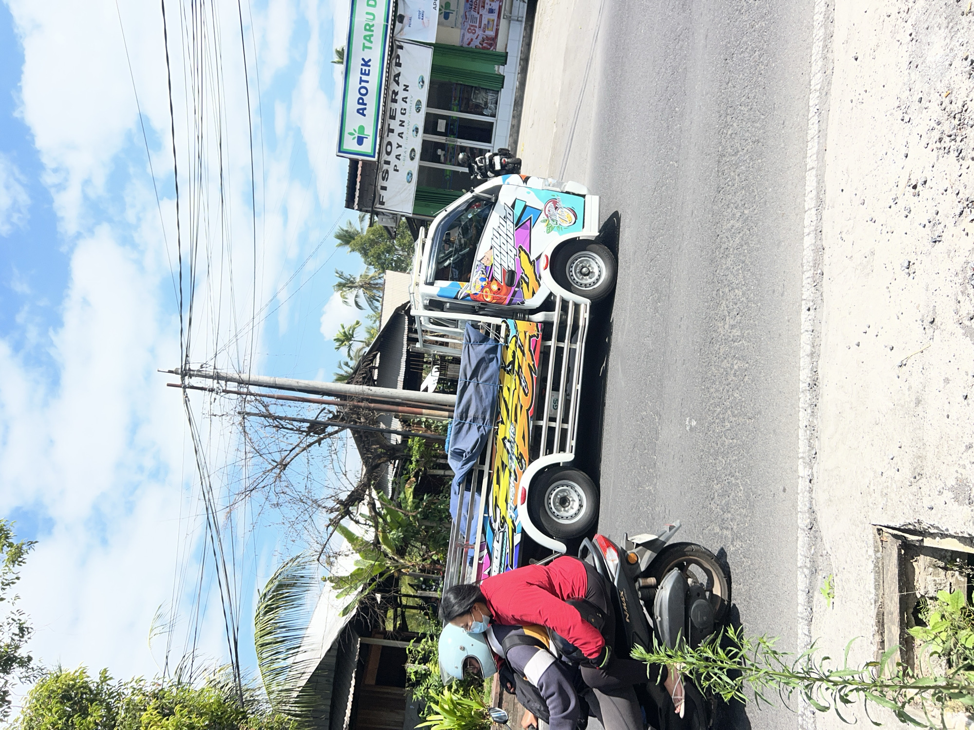 An image taken from the side of a road in Payangan, Indonesia, shows a white flatbed truck with colorful graphics driving past a pharmacy and a physical therapy clinic. Two people are riding on a scooter in the foreground, with the passenger wearing a red jacket and a light blue helmet. The truck is partially covered with blue tarpaulin, and its side features vibrant, stylized artwork. The pharmacy, labeled "APOTEK TARU D," has green and white signage, and the physical therapy clinic next to it is also visible. The scene is set against a backdrop of a bright blue sky with scattered clouds, palm trees, and vegetation. Overhead power lines crisscross the sky, supported by utility poles. The road surface is asphalt, with a concrete shoulder and some sparse greenery on the side.