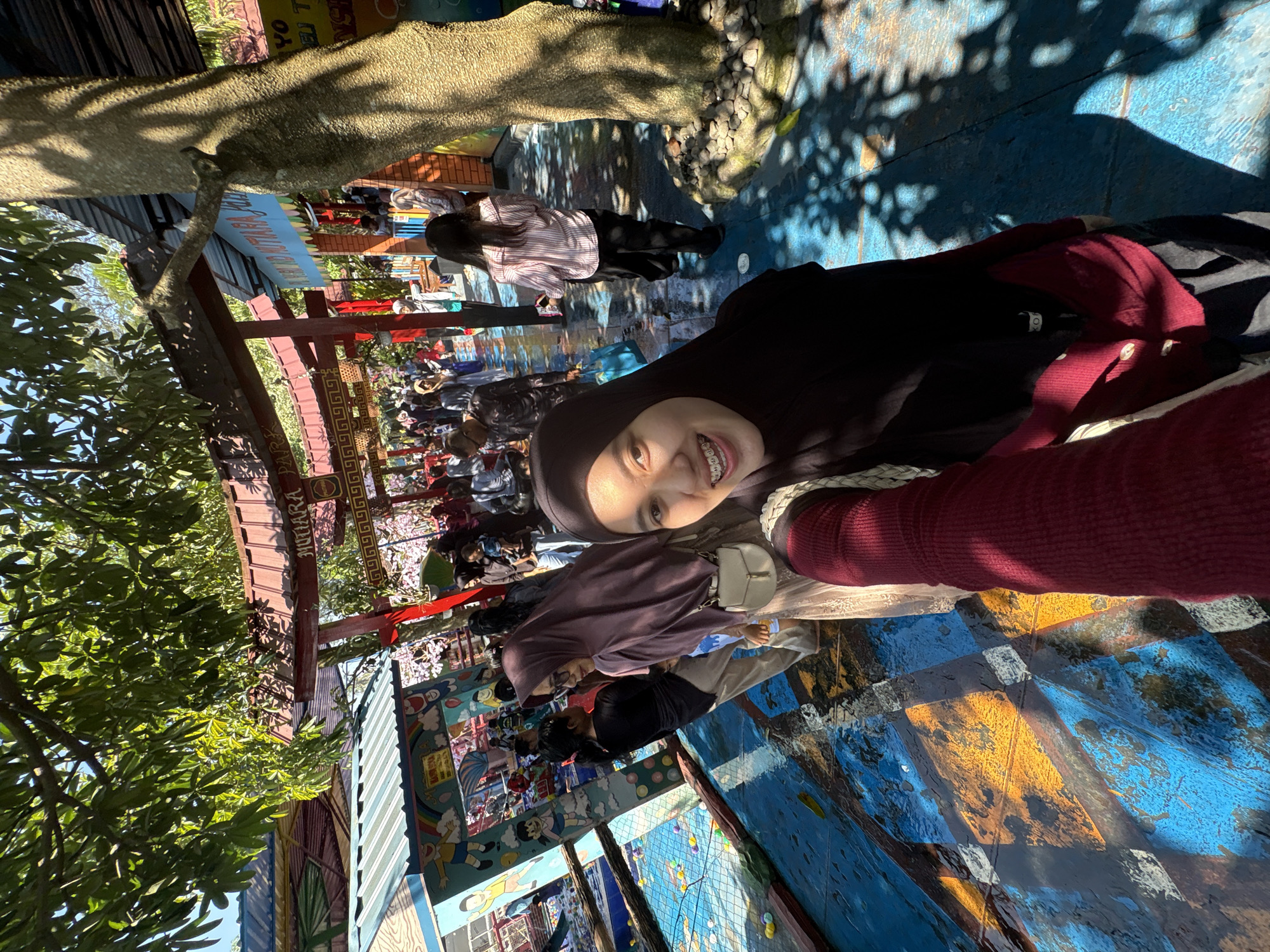 The image captures a bustling scene at the Mutiara Park in Kebonsari, Indonesia. In the foreground, a young woman wearing a black hijab and a maroon cardigan is taking a selfie. Her braces are visible as she smiles at the camera. To her left, another woman in a purple hijab stands, looking away from the camera.
The park features a distinctive red torii gate with the word "MUTIARA" written on it, suggesting a Japanese-inspired theme. Beyond the gate, a crowd of people, many dressed in modest Islamic attire, can be seen enjoying the park. The ground is painted with a vibrant blue and yellow diamond pattern, worn in places, indicating significant use. Colorful murals are visible on a building to the left, depicting cartoon characters. Sunlight filters through the leafy branches of trees, casting dappled shadows on the ground and adding to the lively atmosphere of this recreational space.