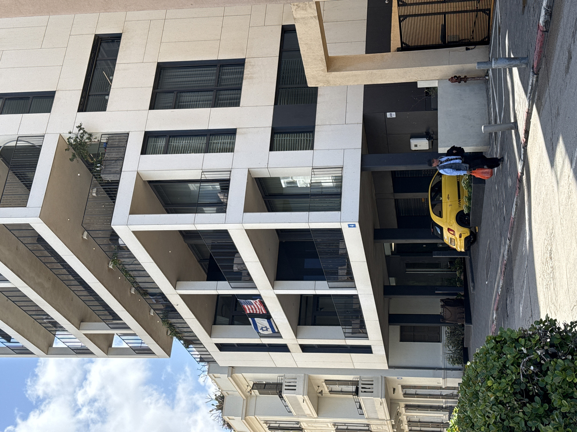 The image shows a street scene in Tel Aviv, Israel, near a modern building with distinctive architectural features. The building is characterized by its white facade, rectangular modules, and recessed balconies with metal railings. An American and Israeli flag are displayed from one of the balconies.
In the foreground, a person is walking alongside a yellow car parked near the building's entrance. The street is paved, and there are bushes and trees along the sidewalk. The lighting suggests it is daytime with bright sunlight casting shadows.