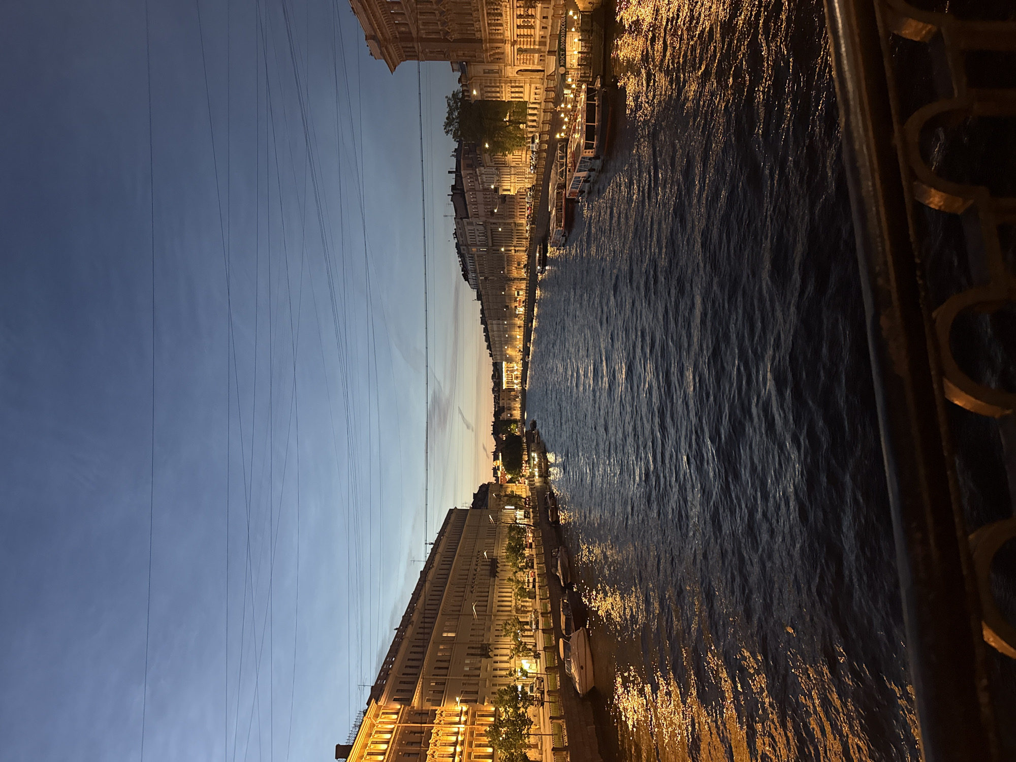 This is a nighttime photograph of a wide canal in Saint Petersburg, Russia. The canal is flanked by ornate, multi-story buildings illuminated by warm, golden lights from windows and street lamps. Several boats are moored along the canal banks, some appearing to be tour boats. The water of the canal reflects the lights from the buildings, creating shimmering patterns on the surface. The sky above is a deep twilight blue, with faint wisps of clouds and several parallel overhead wires crossing the frame. The perspective is from a bridge, with a dark metal railing in the foreground. The overall scene depicts a serene and atmospheric urban landscape at dusk or early night.
