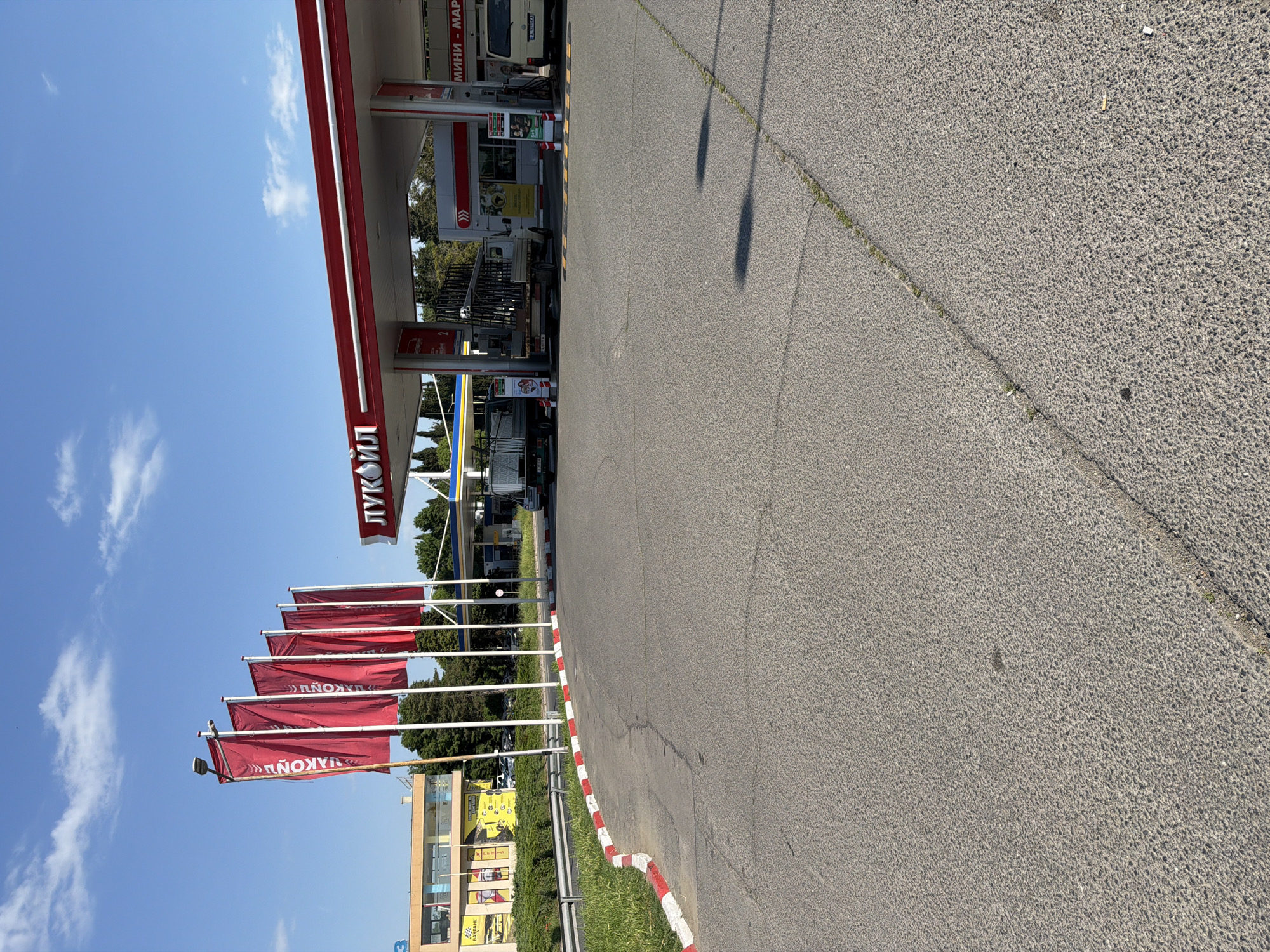 The image shows a Lukoil gas station in Burgas, Bulgaria. Several red flags with the Lukoil logo are displayed in front of the station. The gas station has multiple pumps, and a truck is parked at one of them. The sky is clear with some scattered clouds. A road runs alongside the gas station, with a red and white striped curb. A building is visible in the background. The photo was taken during daylight.