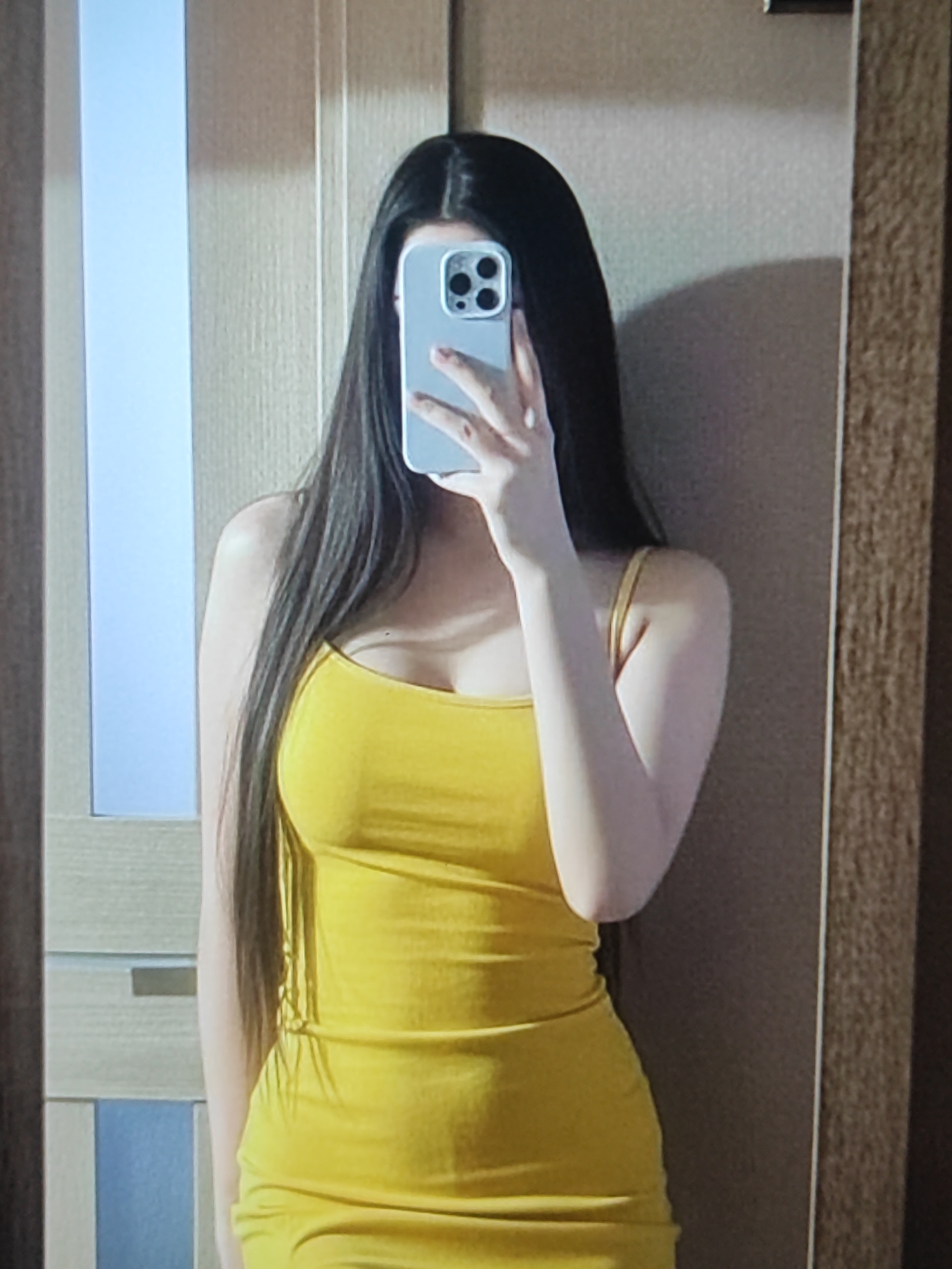 The image shows a person taking a mirror selfie. The person has long, dark hair and is wearing a yellow spaghetti strap dress. They are holding a smartphone up to take the picture, obscuring their face. The background appears to be an indoor setting, possibly a room in a home, with a wooden door and a textured wall visible. There are no discernible location cues suggesting Fergana City, Uzbekistan. The lighting suggests it is daytime.