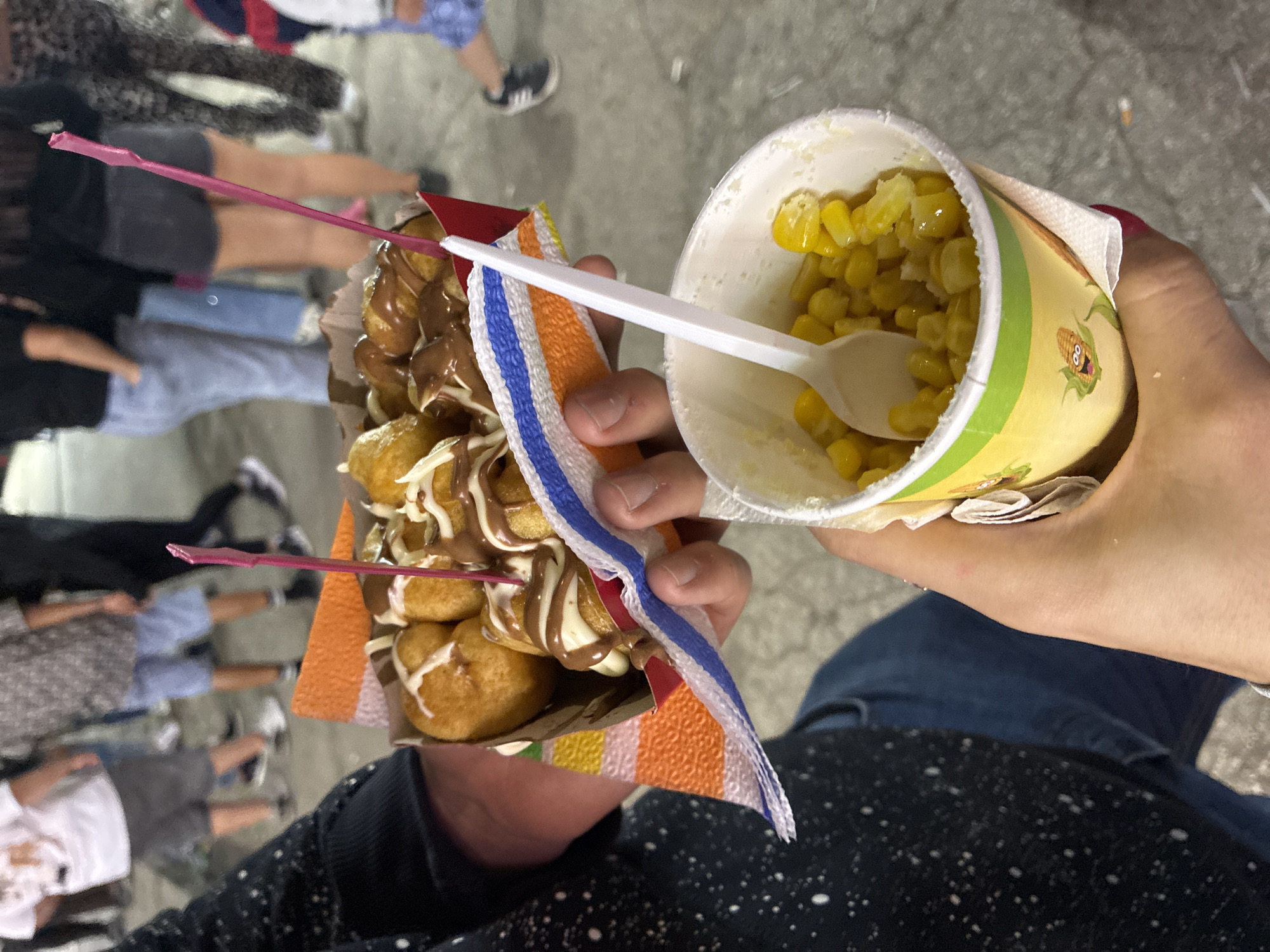 In a dimly lit outdoor setting, likely a fair or market, a person is holding two food items. In their left hand, they grip a red cardboard container filled with golden-brown, fried dough balls drizzled with white and chocolate sauces, skewered by a pink toothpick. To the right, their right hand holds a white paper cup containing yellow corn kernels, with a white plastic spoon resting inside. In the background, blurred figures of people in casual attire are visible, suggesting a public gathering. The ground appears to be paved, possibly concrete. The location is the city of Iskar, Bulgaria.