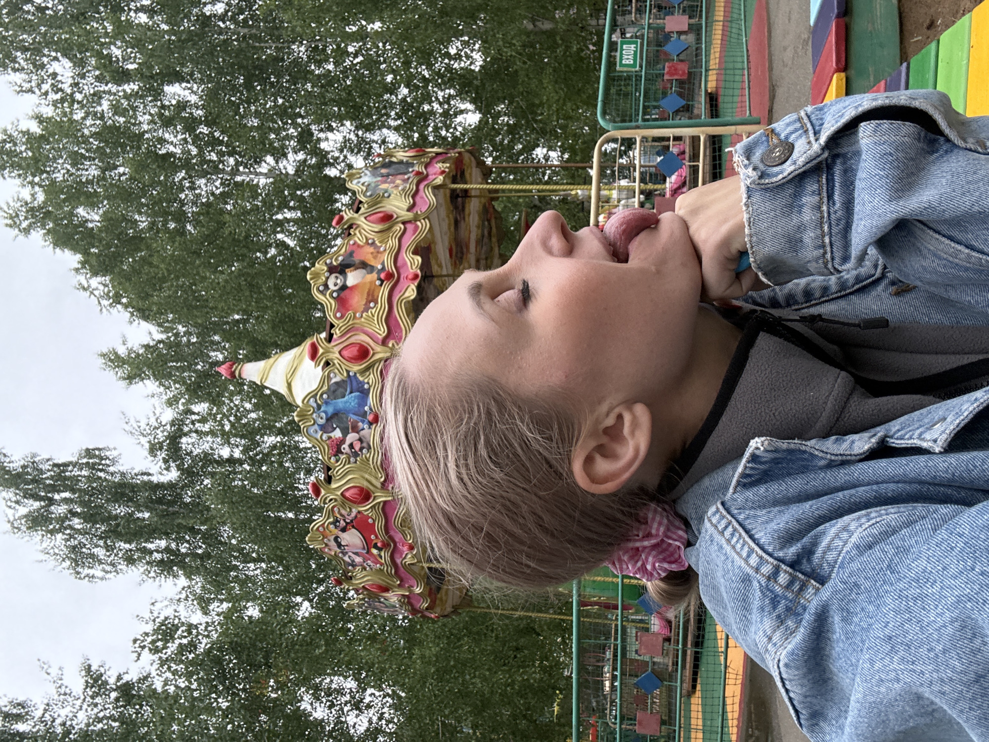 A woman with light blonde hair and fair skin is photographed in an outdoor setting in the city of Chaikovsky, Russia. The woman, wearing a denim jacket and grey fleece, has her eyes closed and her tongue extended toward her fist. In the background, a carousel with cartoon characters is visible, along with trees and some colorful play structures.