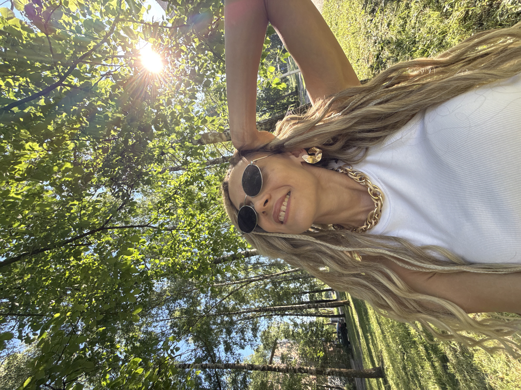 A woman with long, blonde dreadlocks and a white tank top poses for a selfie outdoors. She is wearing large, gold hoop earrings and a chunky gold chain necklace. Sunglasses shield her eyes as she looks up and to the right, a slight smile on her face. Her right arm is raised, with her hand resting on the back of her head. She is in a park-like setting with trees and green grass. The sun is shining brightly through the leaves of a tree, creating a lens flare effect. In the background, there is a building and a dark-colored car parked on a street.