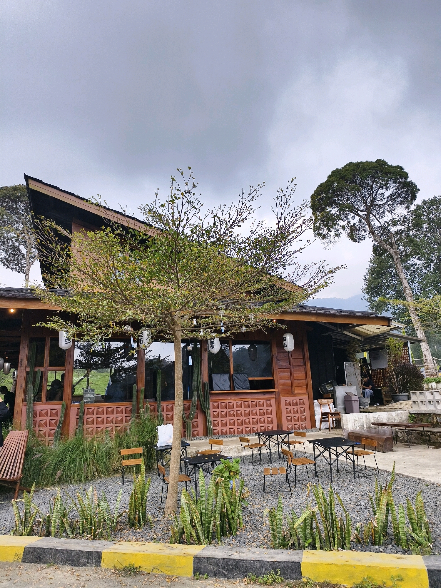 The image depicts the exterior of a wooden building, likely a cafe or restaurant, situated in Patengan, Indonesia. The building features a distinctive asymmetrical roofline and large glass windows revealing the interior. In front of the building, outdoor seating arrangements are visible, including tables and chairs scattered across a gravel-covered area. Various plants, including succulents and tall grasses, are planted around the seating area, adding greenery to the scene. A tree with sparse green foliage stands prominently in the foreground, partially obscuring the building. The sky is overcast, suggesting a cloudy day. In the background, trees and a hillside are visible, indicating a natural or semi-rural setting. Several people are present inside and outside the building, engaged in various activities. The overall impression is of a casual, outdoor dining or relaxation space within a scenic environment.