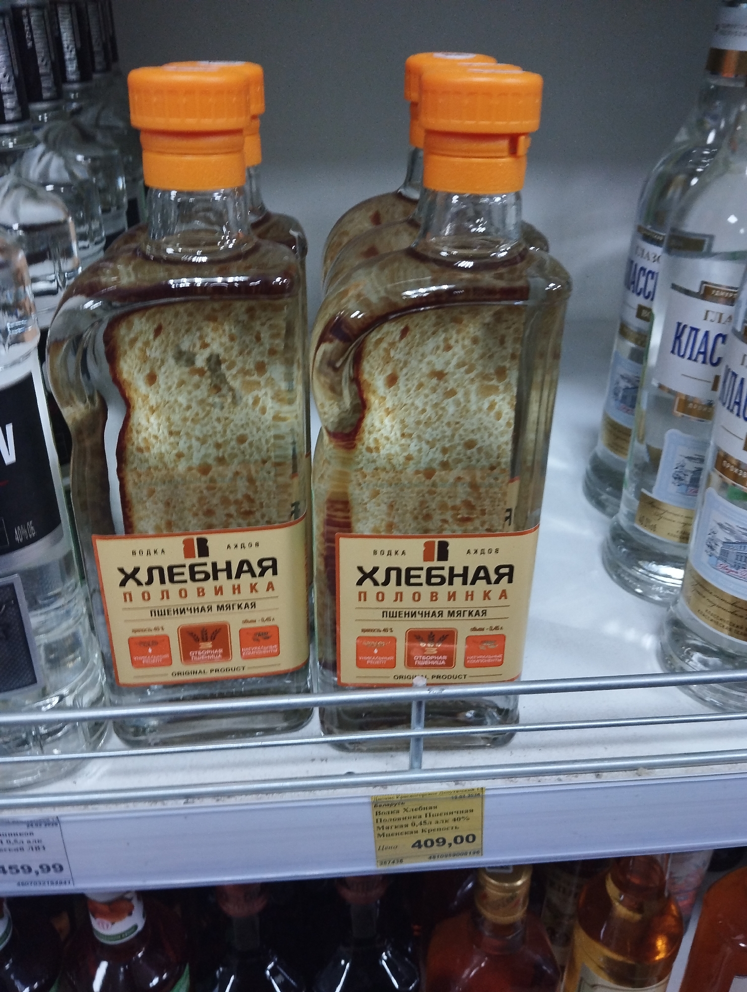 The image shows two bottles of "Khlebnaya Polovinka" vodka on a shelf in a store in Krasnogorskoye, Russia. The bottles are shaped like bread loaves and are positioned side-by-side, displaying their labels prominently. The vodka label states it is made from "wheat," and the bottle has an orange cap. Other bottles of vodka and various alcoholic beverages are on the shelf nearby. The price tag below the bottles indicates a price of 409.00 (currency unspecified). Another price tag indicates a price of 459.99.