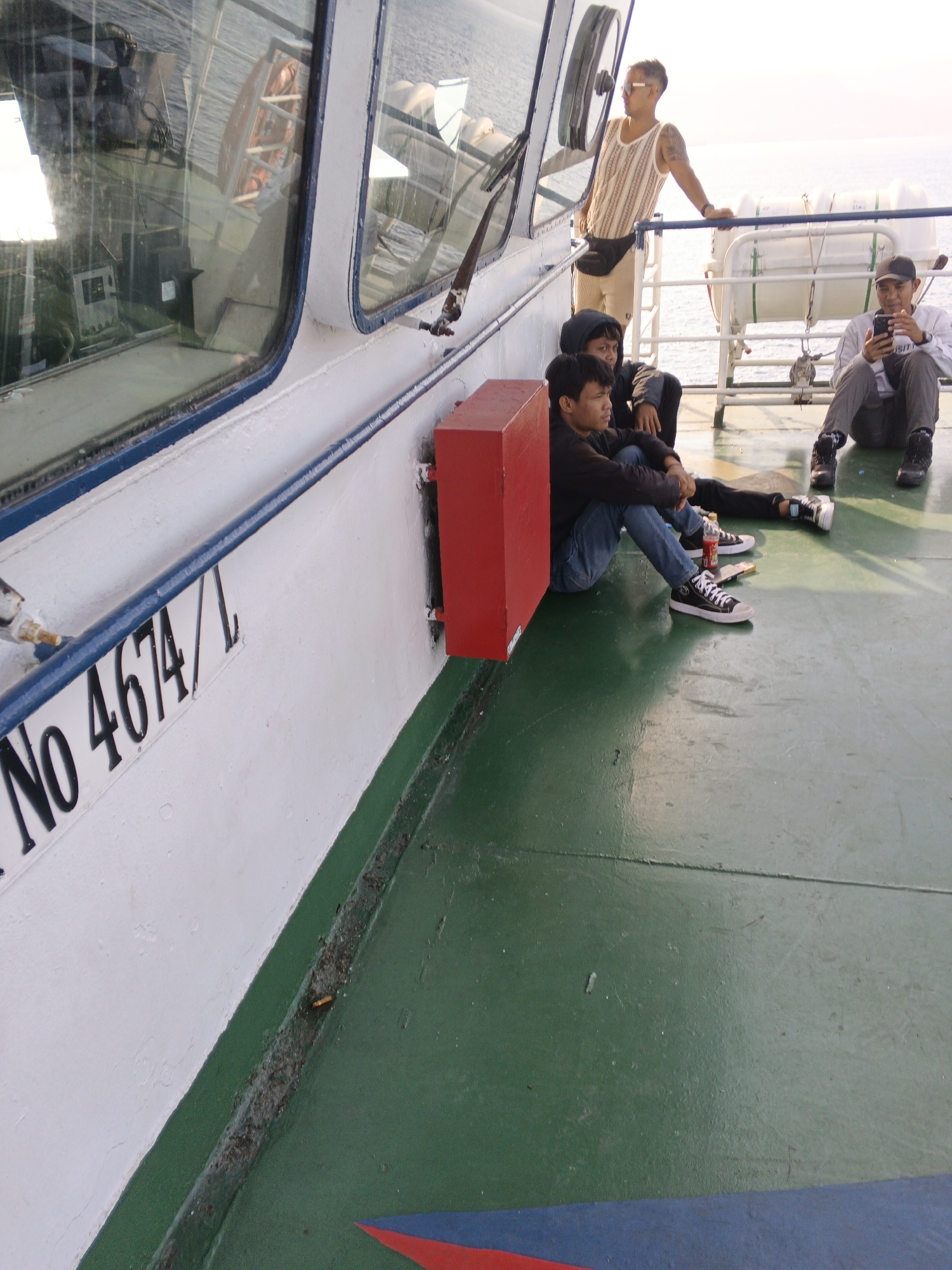 In an outdoor setting on a boat deck, four individuals are present. One man, wearing sunglasses, a sleeveless striped shirt, and khaki shorts with a black fanny pack, leans against a railing. Another man, wearing a light-colored long-sleeved shirt and dark pants, is seated and looking at his phone. Two other young men are seated on the green deck. One, in a black hooded jacket and jeans, sits with his legs extended, while the other, wearing a dark long-sleeved shirt and blue jeans, is seated with his knees drawn up. A red storage box is positioned to the left of the seated individuals. The deck of the boat is painted green, with a white section bearing the black lettering "No 4674/L". The background shows the ocean and life rafts. The scene appears to be in Unknown, Indonesia.