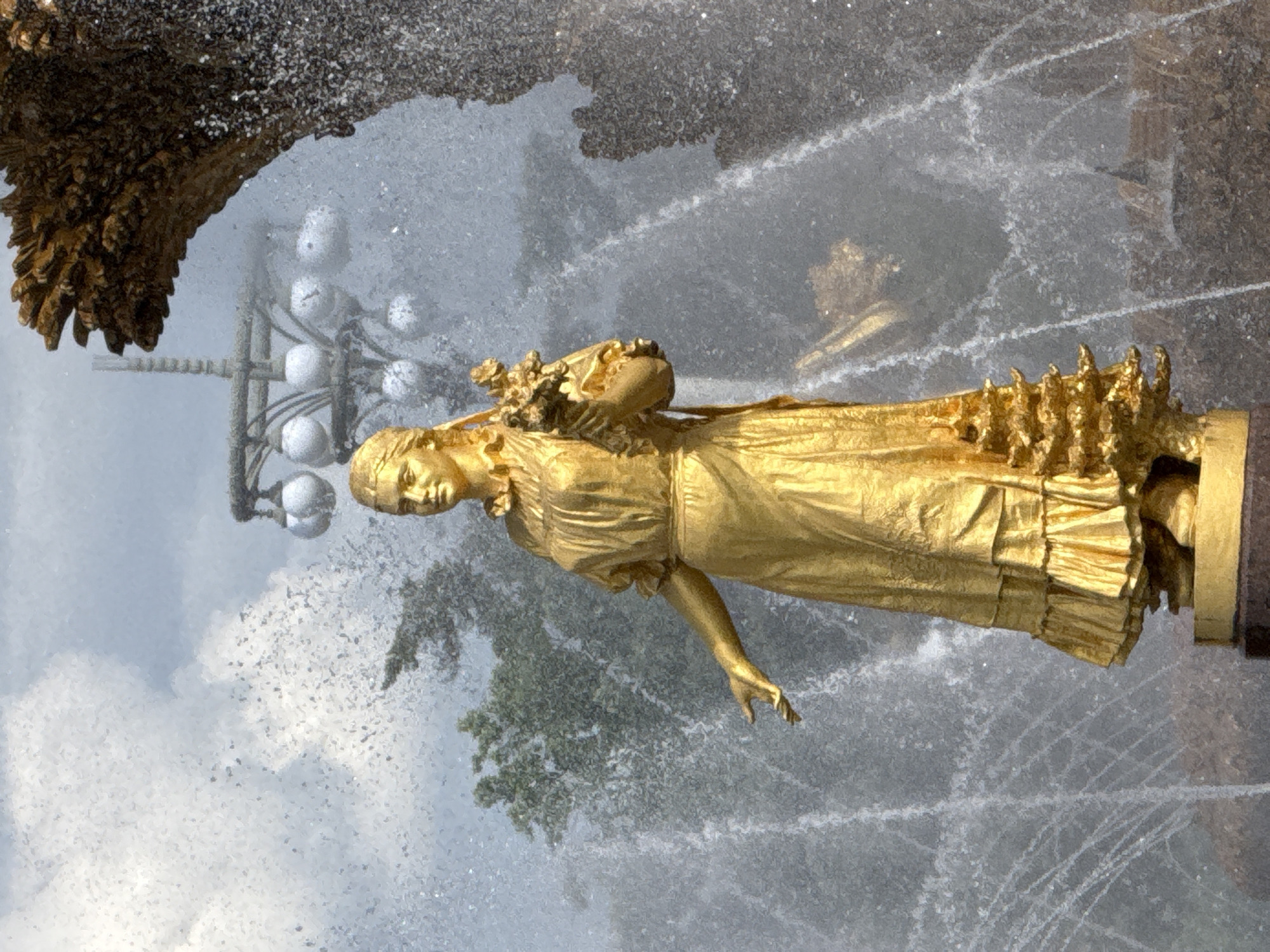 This media file features a golden statue of a woman, partially obscured by water from a fountain. The statue depicts a woman in a flowing gown, holding a bouquet of flowers. Her left arm is extended, and her head is tilted slightly. The statue is positioned in what appears to be a park or public garden, with trees visible in the background. The water from the fountain creates a shimmering effect, adding to the visual appeal of the scene. The statue's golden hue suggests it is made of bronze or gilded metal. The overall mood is serene and artistic, with the water adding a dynamic element to the static sculpture. The scene is illuminated by natural light, suggesting it is daytime. The location context provided is Moscow, Russia.