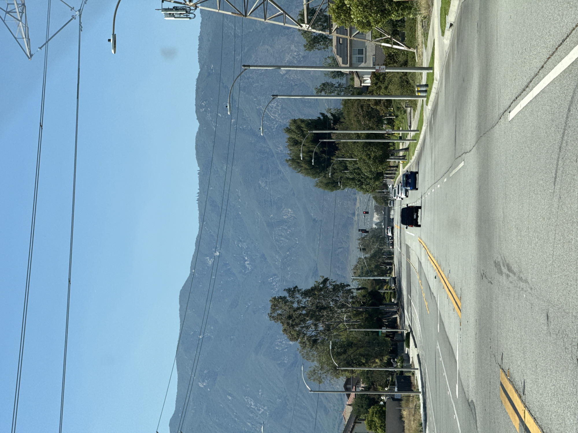 The image depicts a street scene in Fontana, California, USA. A wide, multi-lane asphalt road stretches into the distance, with yellow and white lane markings visible. Several cars are present on the road, with a black SUV in the foreground and other vehicles further ahead. The road is lined with streetlights and mature trees. In the background, a large mountain range dominates the horizon under a clear blue sky. Utility poles and power lines are also visible, as is a tall metal tower on the right side of the image. Residential buildings with pitched roofs are situated along the sides of the road. The overall impression is of a suburban or semi-urban environment with a natural landscape backdrop.