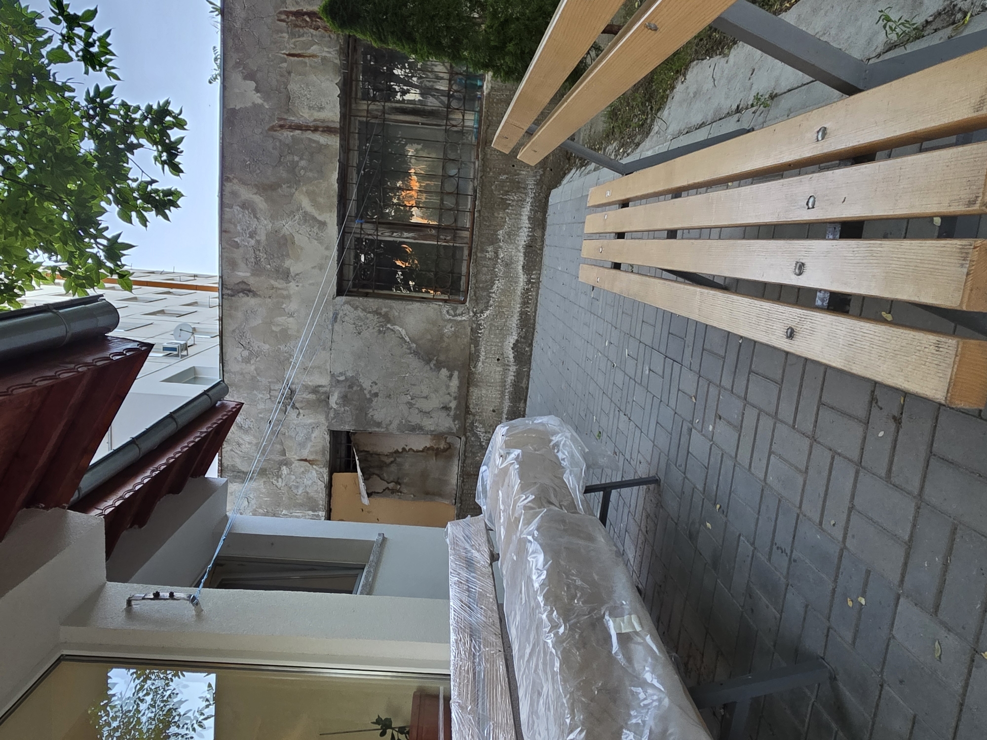 The image shows an outdoor scene in Pleven, Bulgaria. A section of a weathered building with a barred window is visible on the left. Next to it is a newer building with a red-tiled roof. Two benches stand on a gray brick pathway. One bench is empty and the other is covered in clear plastic wrap. A green tree is seen in the top center of the image.