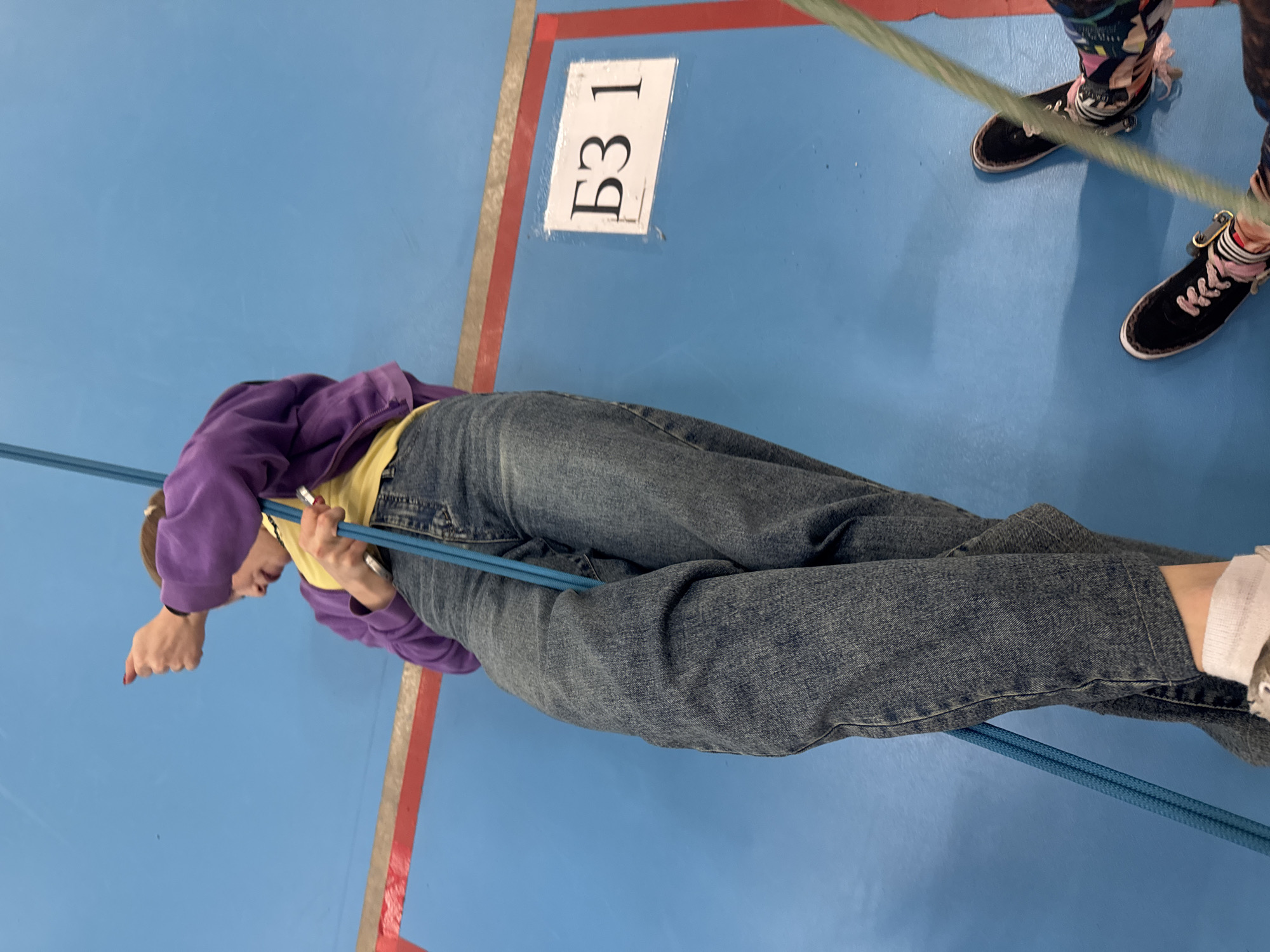 The image depicts a person in jeans and a purple jacket lying on a blue surface with red and white markings. The surface appears to be the floor of an indoor space. The person is holding onto a blue rope. There is a white sign with the text "БЗ 1" on the floor. Another person's legs and shoes are visible in the upper right corner. The location is possibly in Usolye, Russia.