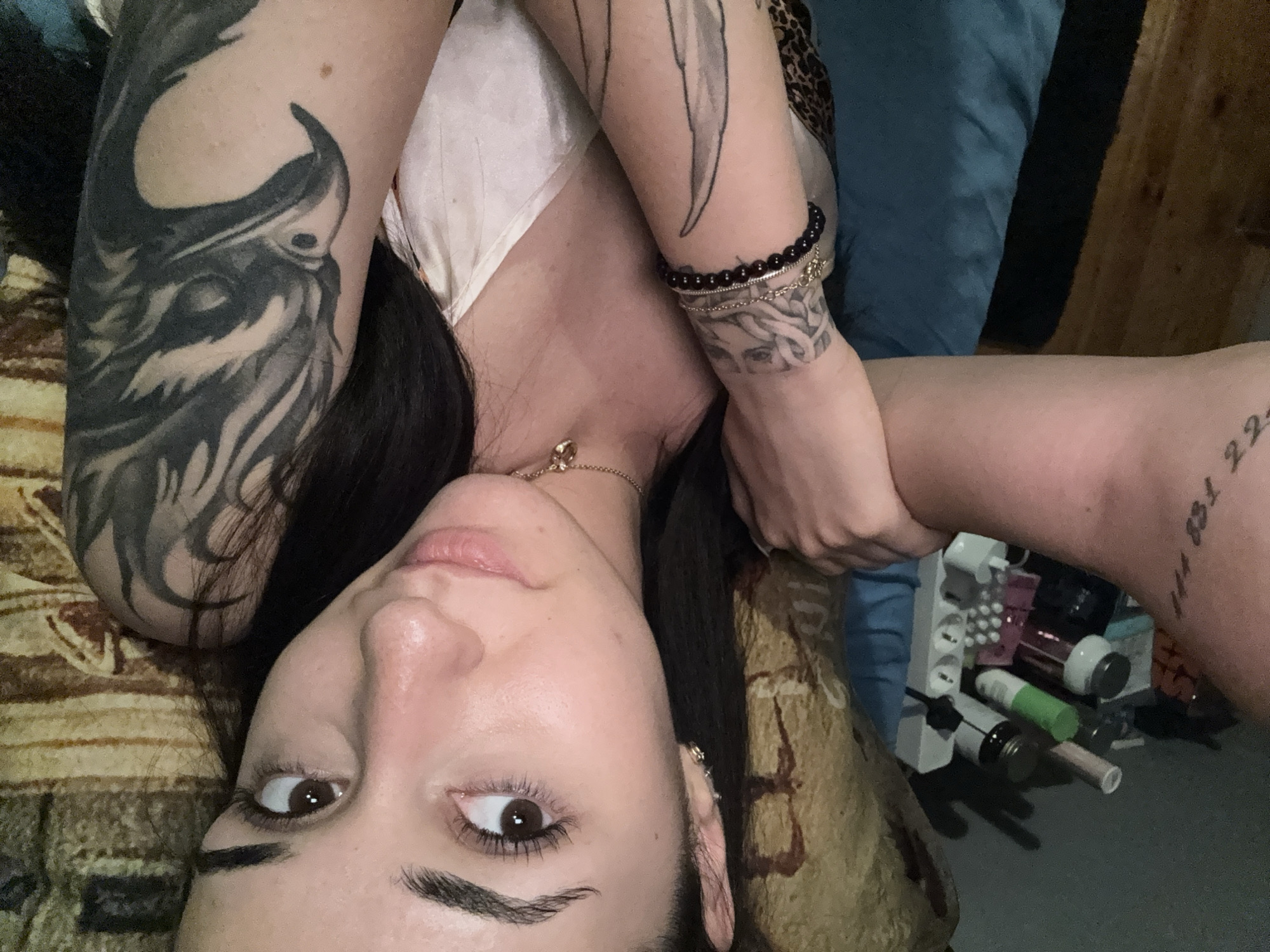 A young woman is lying on a bed, looking up at the camera. She has dark hair and tattoos on her arms. She is wearing a light-colored top. In the background, there is a blue blanket and a shelf filled with various items, including bottles and containers. The room appears to be a bedroom. There is no indication of the city of Shumen, Bulgaria, or any other specific location within the image.
