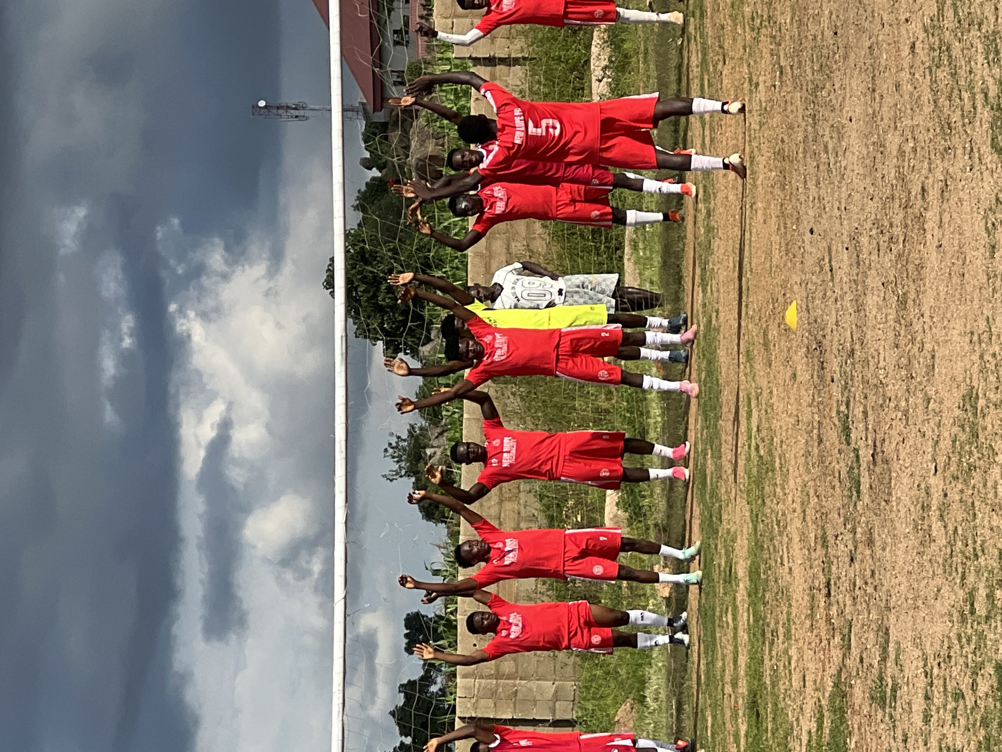 In Jos, Nigeria, a group of young men wearing red soccer uniforms and white socks are seen standing on a dirt soccer field, facing a net. Their arms are raised in unison, suggesting a moment of team unity or a pre-game ritual. One individual in the background wears a white jersey with the number "09" visible. The sky above is overcast with dark, heavy clouds, hinting at impending rain or stormy weather. The surrounding environment includes a stone wall behind the players, some trees, and a distant tower with antennas, likely a telecommunication mast.