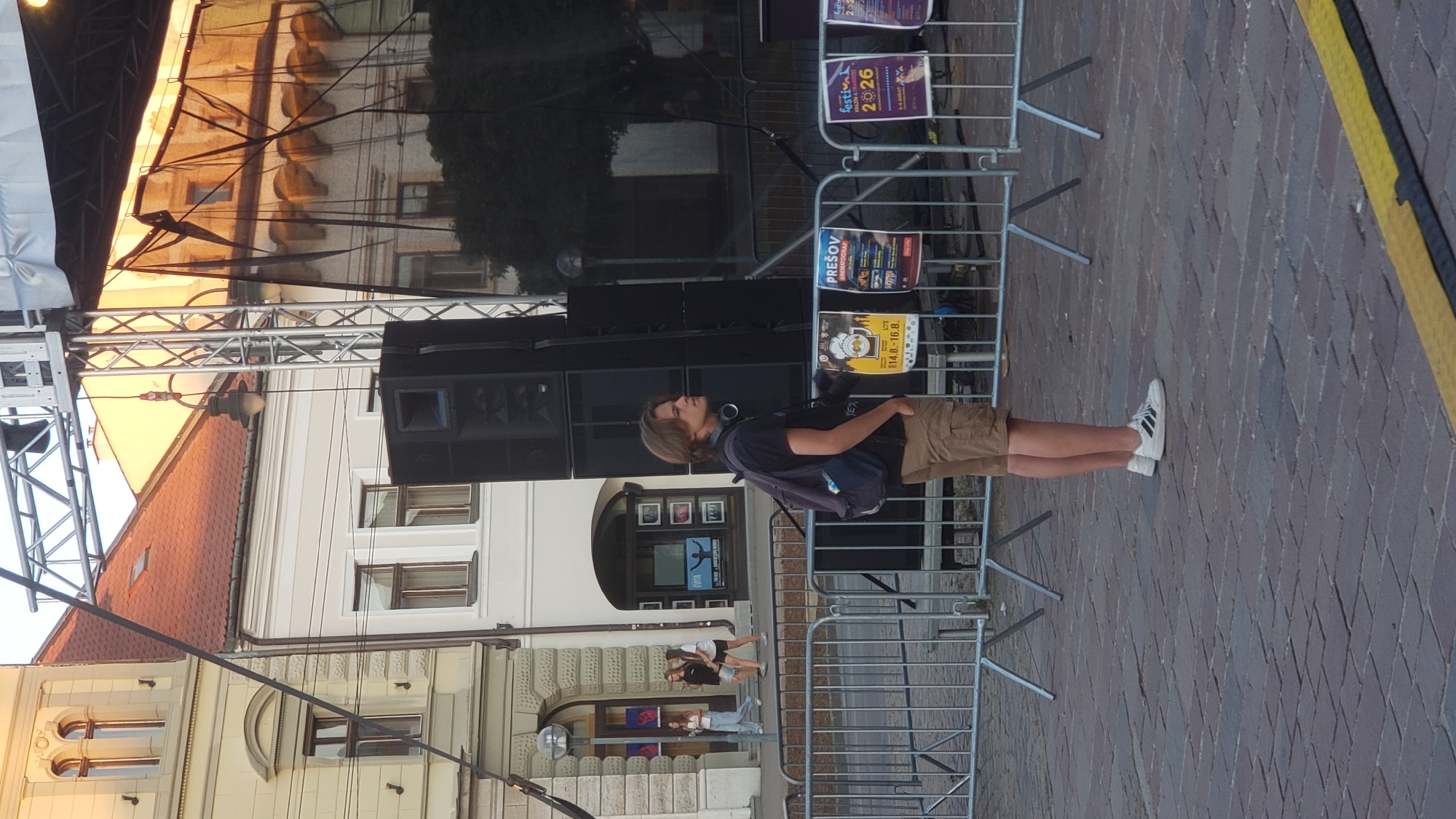 A young person with light brown hair stands on a cobblestone street in Prešov, Slovakia. They are wearing a dark t-shirt, tan cargo shorts, and white Adidas sneakers with black stripes. A dark backpack with a blue pouch is slung over their right shoulder. The person is facing right, looking slightly upwards. Behind them is a large sound system stack, a metal barrier, and several posters. One poster visible in the foreground has the text "FESTIVAL" and the number "2026" in large print. Another poster reads "PREŠOV KINEMATOGRAF". In the background, there are buildings with red tiled roofs and windows, some with ornate architectural details. Sunlight casts a warm glow on the buildings. A black mesh curtain hangs in front of a stage structure. The scene appears to be set up for an outdoor event or festival.
