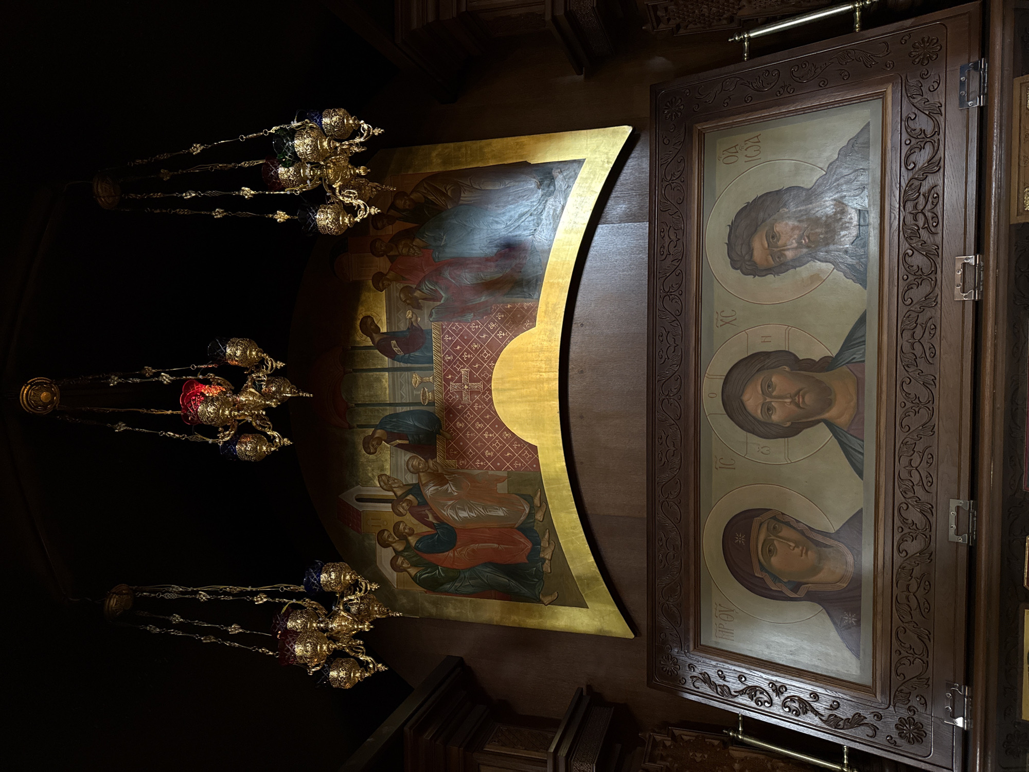 The image depicts the interior of a religious space, likely a church, featuring Byzantine-style art and liturgical objects. Three hanging lamps, made of gold-colored metal and adorned with colored glass elements, are visible on the left. A curved painting, framed with gold, is centrally located. It depicts several figures seated around a table. To the right of this painting is a wooden cabinet featuring an icon with three portraits: an older bearded man, a man with short hair, and a woman wearing a head covering. The scene suggests a sacred or devotional setting, with the artistic style being characteristic of Eastern Orthodox religious art. There is no information present in the image about the location being Nizhny Novgorod, Russia.