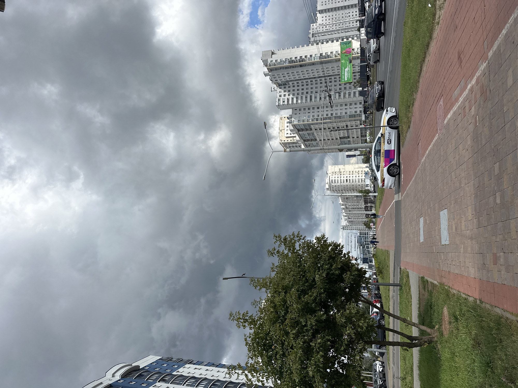 The image depicts a cloudy day in a modern urban environment, likely in Minsk, Belarus, as suggested by the signage. In the foreground, a red-brick pedestrian walkway runs along the side of a multi-lane road. To the left of the walkway is a patch of green grass with a young tree and a manhole cover.
Across the road, a stream of vehicles is visible. A white "wb taxi" is in the left lane, and behind it are other cars and a dark SUV on the right. The road is lined with several tall, contemporary apartment buildings. On the far right, a billboard with green accents and text in Cyrillic can be seen. The sky is dominated by heavy, dark grey clouds, with a small patch of blue sky visible in the upper right corner. A building with blue and white vertical stripes is partially visible on the far left. The scene suggests a typical day in a developed residential area of a city, with an impending sense of possible rain due to the sky conditions.