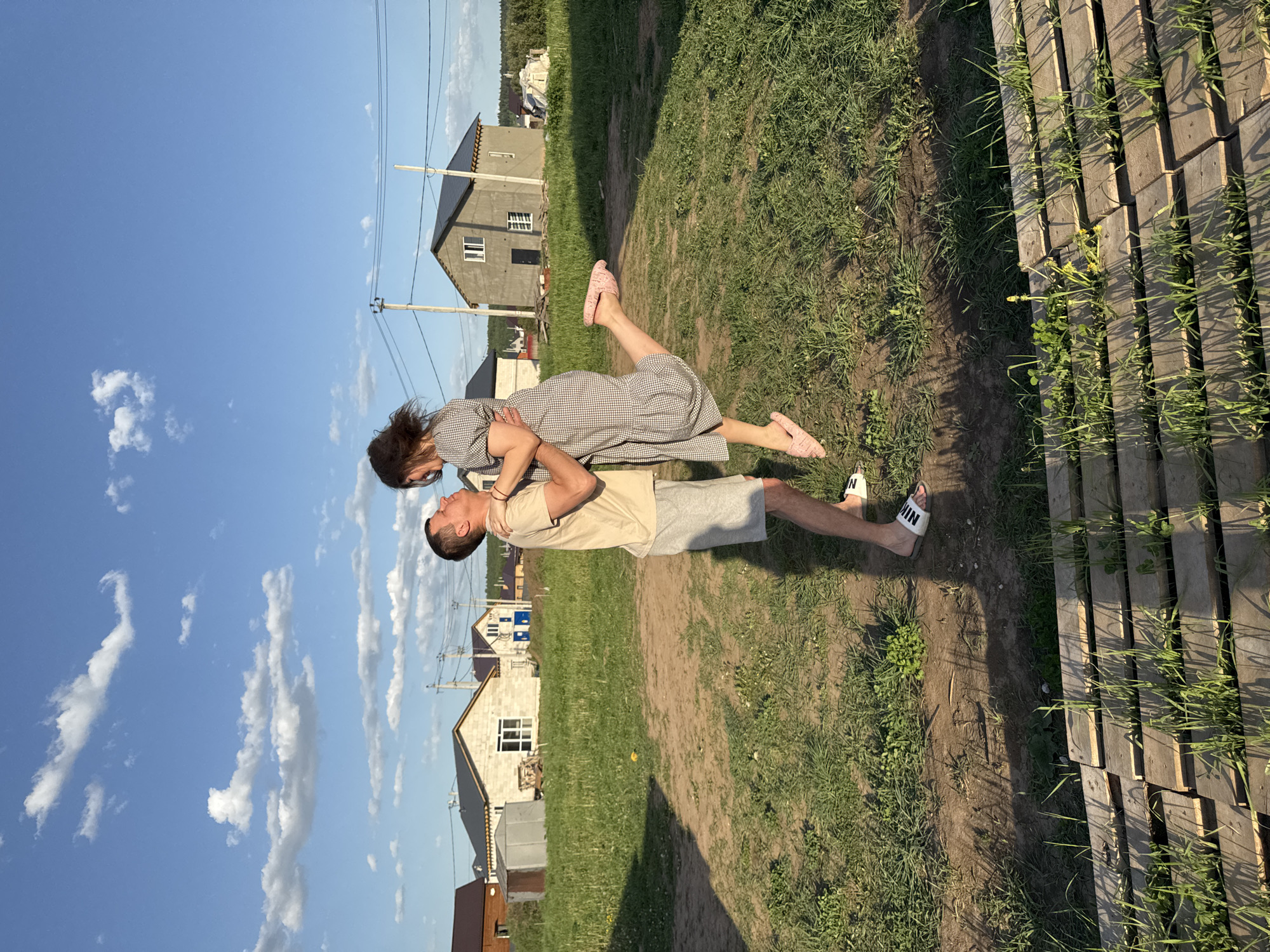 A man and a woman are pictured outdoors in Ферма, Russia. The man is holding the woman in the air. She is wearing a gray and white checkered dress. The man is wearing a tan t-shirt and gray shorts. They are standing on a grassy path with houses in the background. The sky is blue with white clouds. A stack of wooden pallets is to the right of the frame. The man is wearing white sandals with black Nike branding.