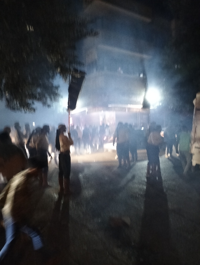 The image depicts a nighttime scene in the city of Homs, Syria, characterized by low light, blur, and atmospheric haze. A crowd of approximately 20-30 individuals is gathered outdoors, predominantly in the mid-ground and background. Artificial light sources illuminate the scene, casting strong beams through visible haze or smoke, and creating bright reflections on the dark, wet ground.
In the mid-ground, slightly to the left of center, an individual stands holding an upright dark-colored flag or banner on a pole. This person wears a light top and dark trousers. Other individuals in the crowd are indistinct due to blur and distance, appearing to be standing or slowly moving. In the immediate foreground, lower left, a person is heavily blurred by motion, suggesting rapid movement or a longer exposure capture of their movement, wearing blue trousers and a light-colored top. Elongated shadows of individuals are cast onto the reflective ground surface.
The background includes a multi-story building, partially visible with some illumination, and silhouetted trees on the left. The overall environment appears to be an urban setting, possibly a street or open area, with a pervasive smoky or foggy atmosphere.