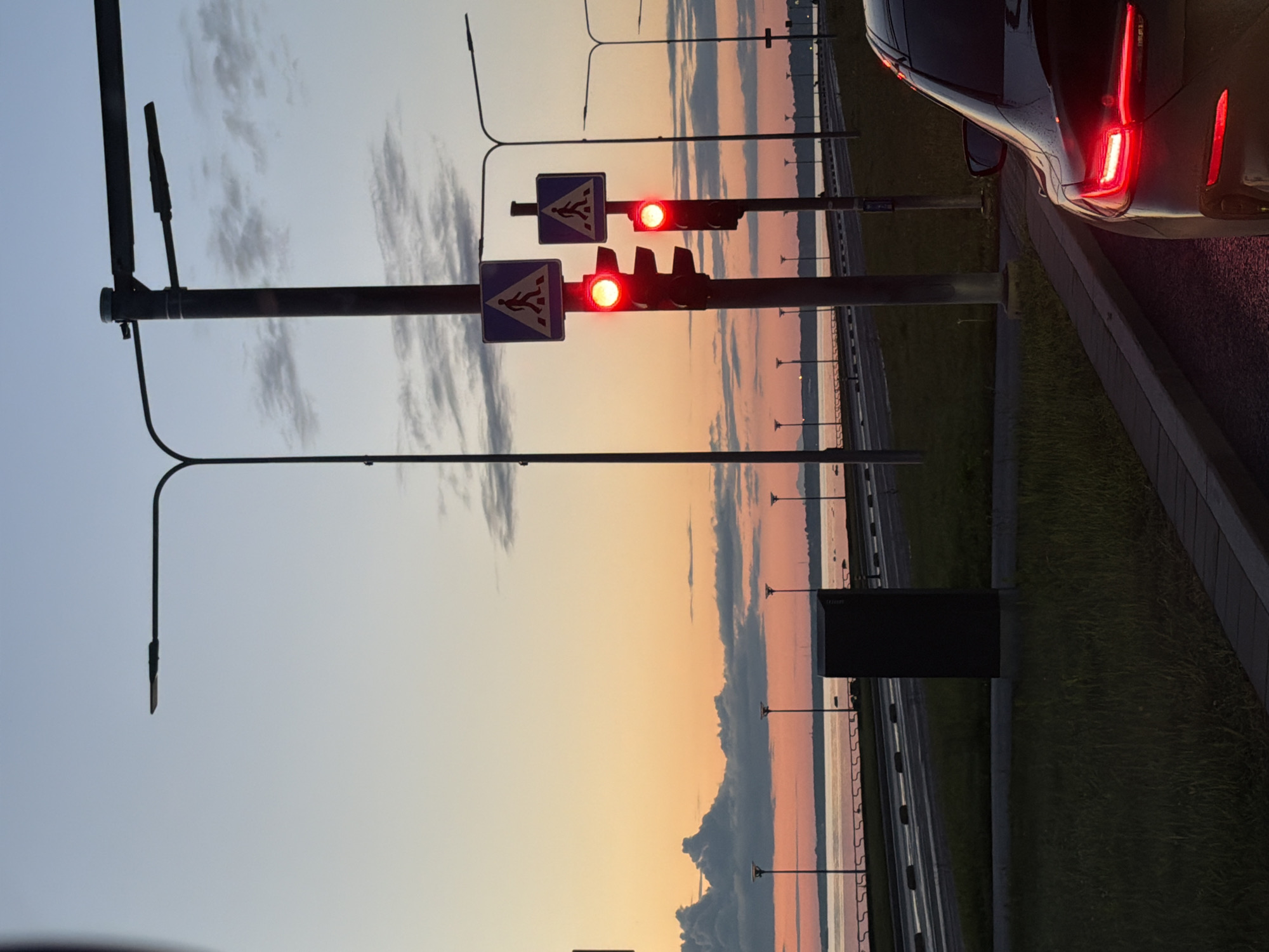 At dusk in Tallinn, Estonia, a black car is stopped at a traffic light, its red taillights illuminated. The traffic light, displaying a red signal, is positioned next to a pedestrian crossing sign. The scene is located by the sea, with a body of water visible in the background. The sky is a gradient of pale blue and orange, with some clouds scattered across it. Streetlights line the road, which appears to be a coastal highway. The overall ambiance is calm and serene, with the natural light of sunset complementing the artificial lights of the city.