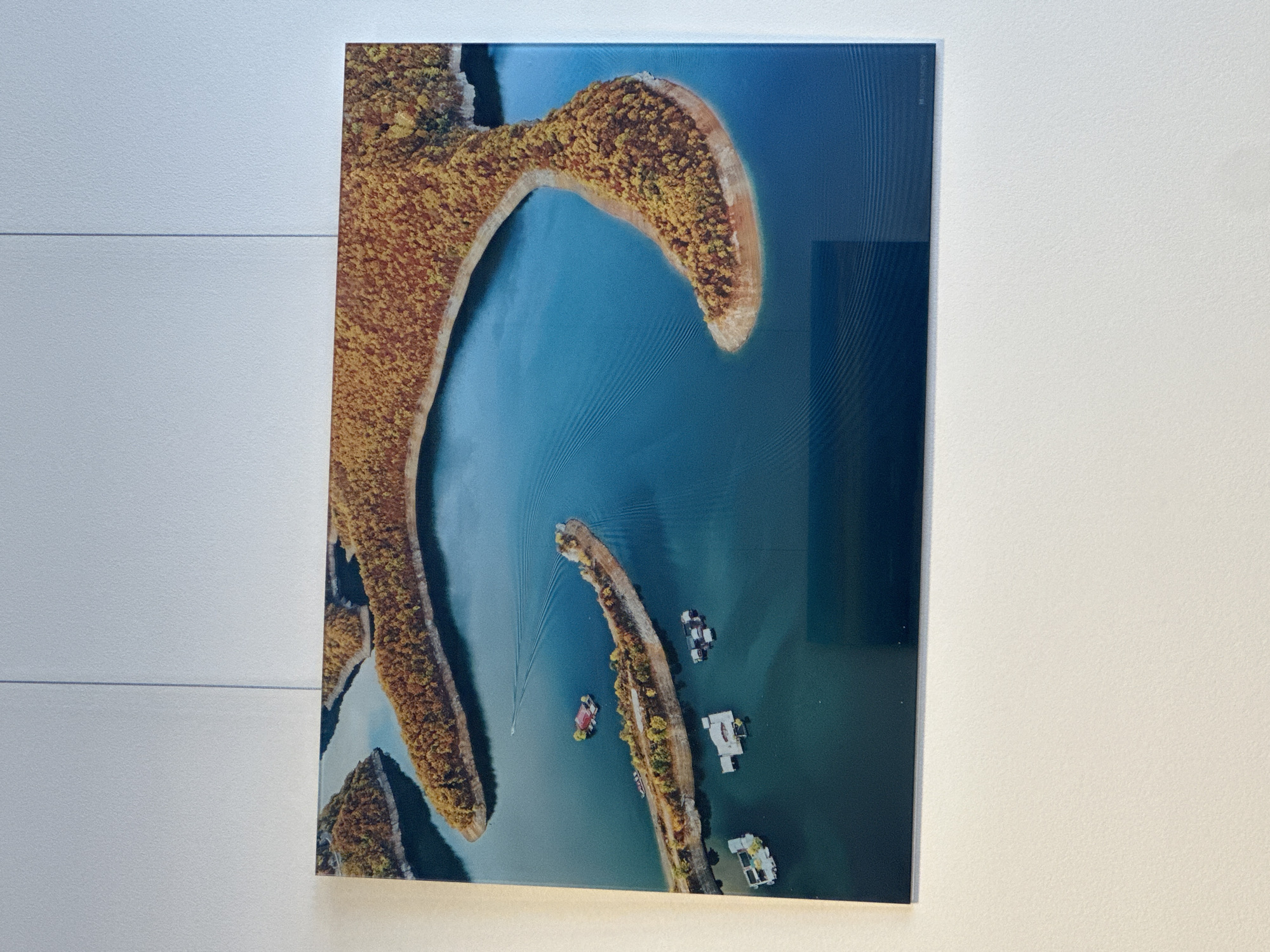 This is an aerial, wide shot of a body of water with several islands covered in autumn foliage. The water is a deep blue, with lighter blue trails indicating the movement of boats. Several structures resembling houseboats or floating homes are anchored in the water near the islands. The largest island forms a crescent shape, with a smaller, elongated island to its left. The image is framed and hanging on a white wall.