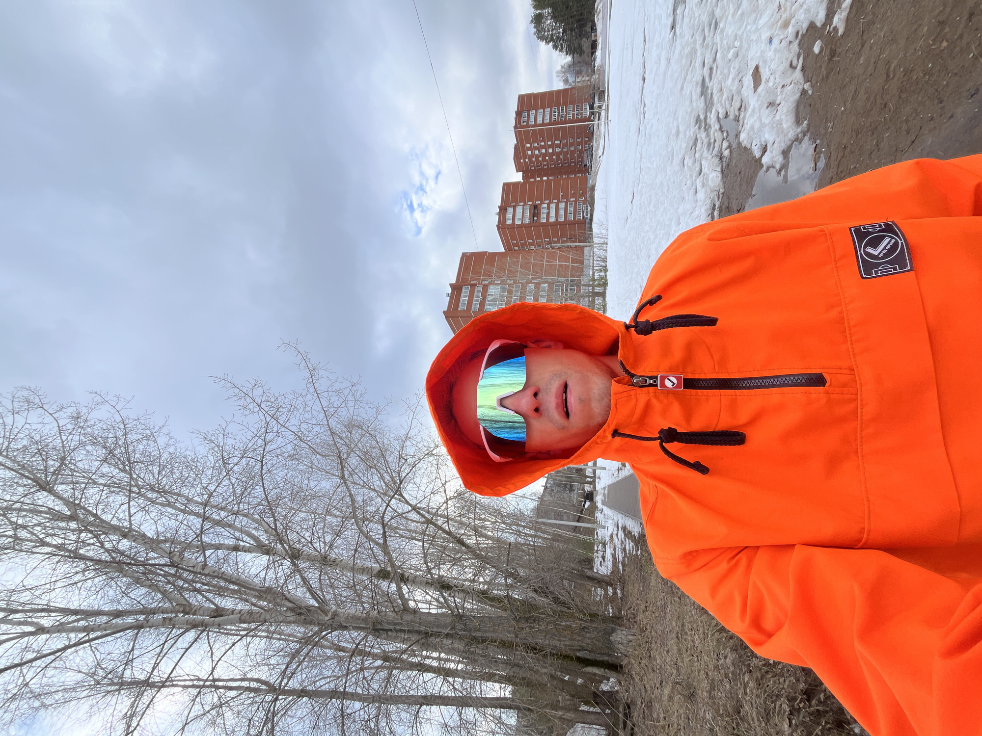 A man wearing an orange hooded jacket and mirrored sunglasses stands outdoors in Chaikovsky, Russia, under a cloudy sky. Buildings are visible in the background, and there is snow on the ground near what appears to be a dirt or gravel path. Leafless trees are in the foreground. The man is looking at the camera and seems to be taking a selfie.