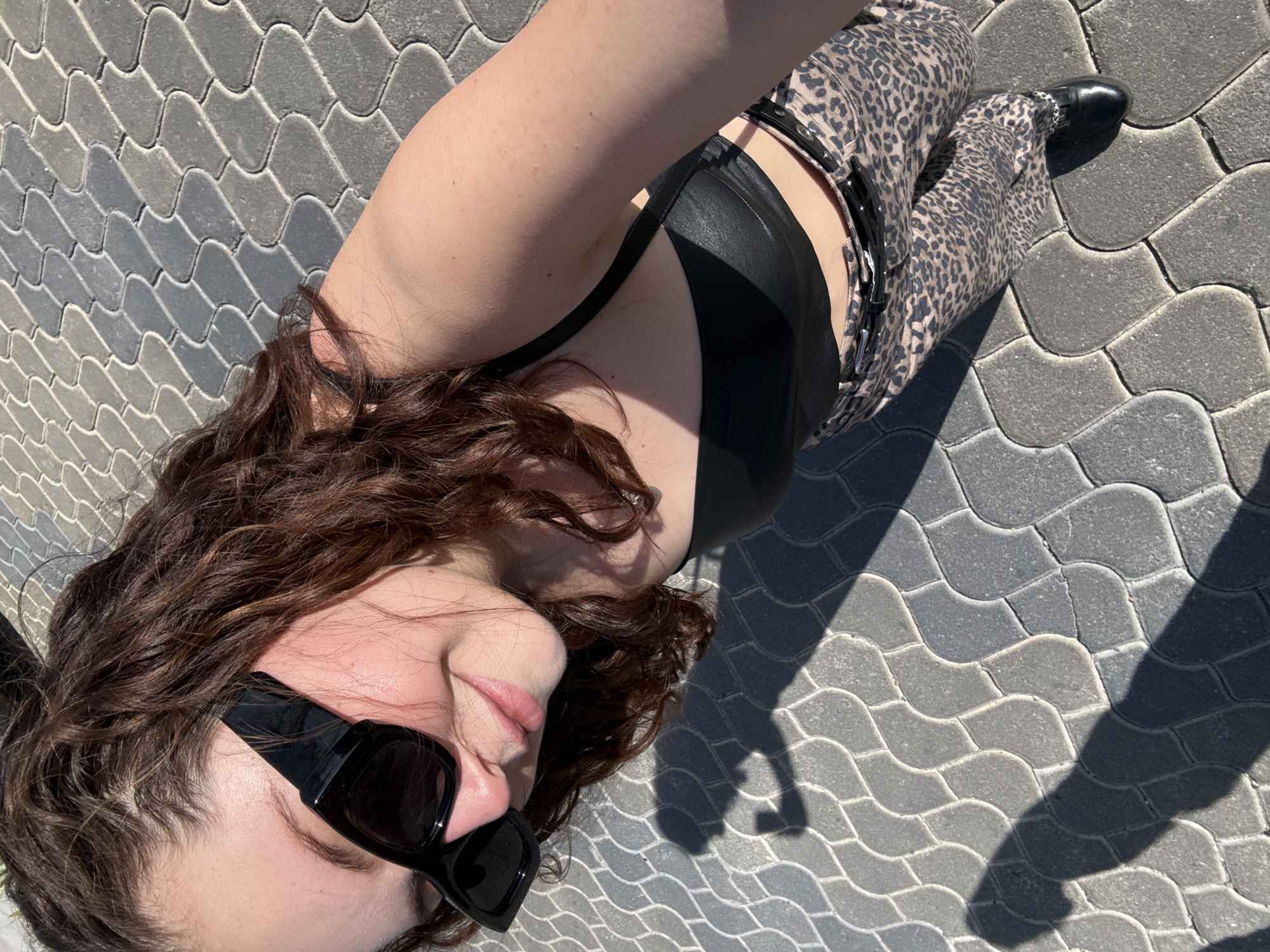 A woman with sunglasses, a black top, and leopard-print pants is lying on a gray paved surface, possibly a sidewalk or patio. The location is Santa Clara Ocoyucan, Mexico. The woman's hair is brown and curly. Her left foot is visible, clad in a black shoe. She appears to be taking a selfie.